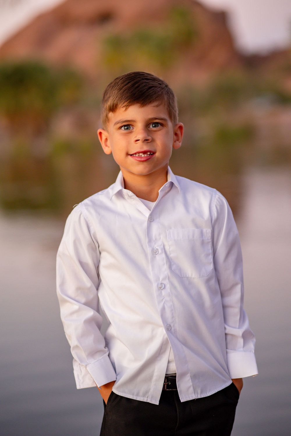 Smiling young boy in white dress shirt posing outdoors near a lake with rocky desert backdrop.