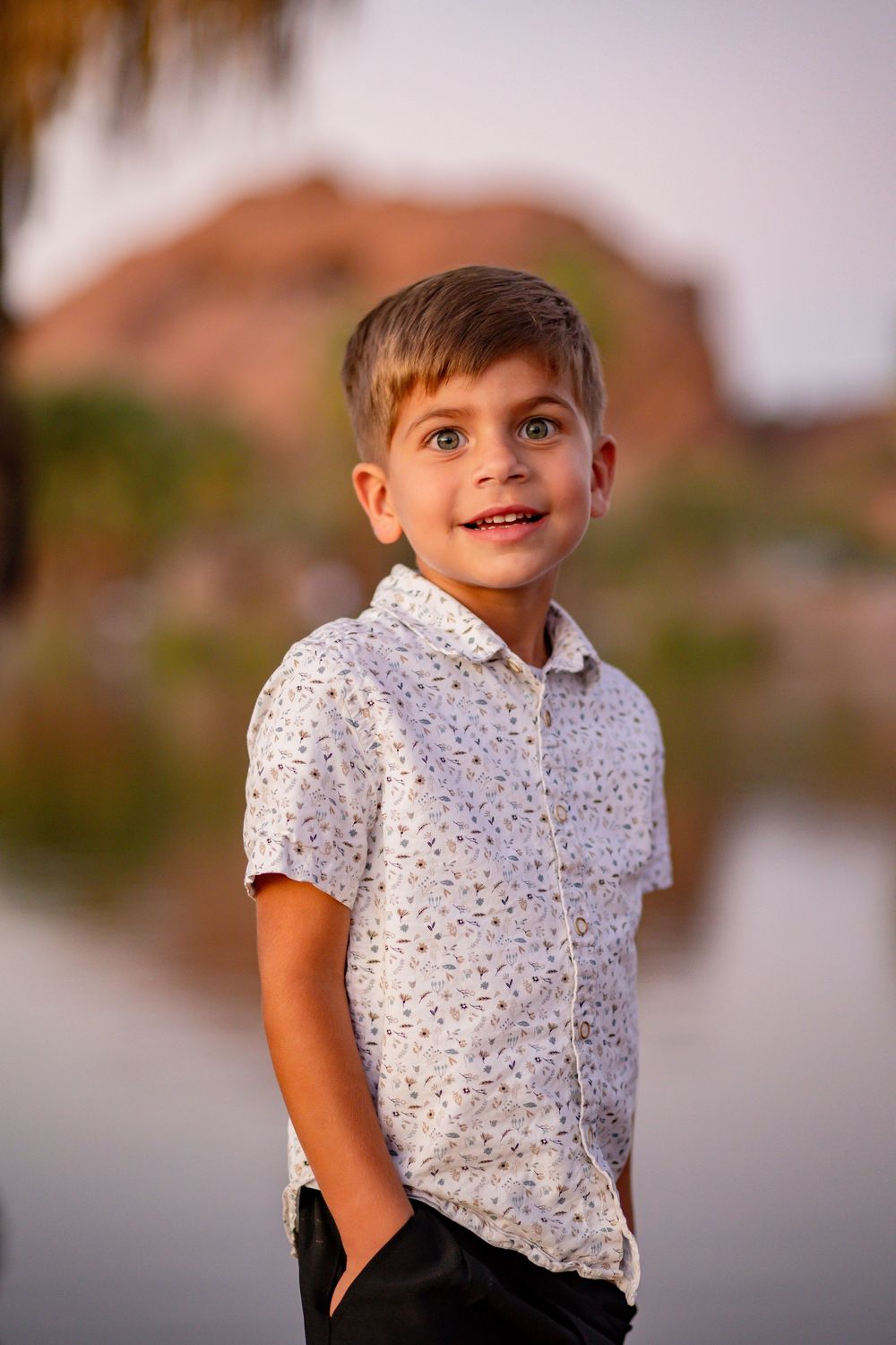 Cheerful young boy in patterned white shirt standing outdoors near a lake with desert mountain background.