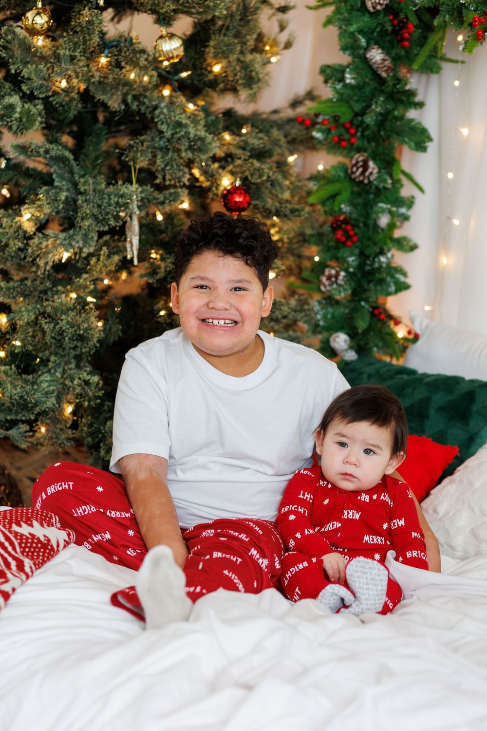 Smiling older brother sitting with baby sibling on white bed in front of decorated Christmas tree.