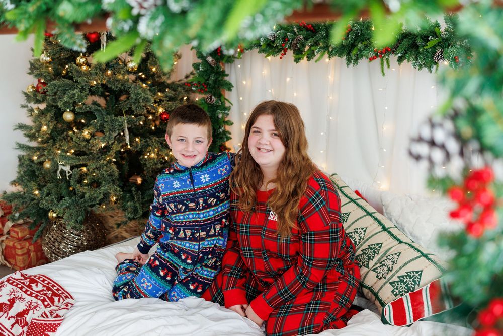 Two siblings in matching Christmas pajamas smiling together on a bed with festive holiday decorations.