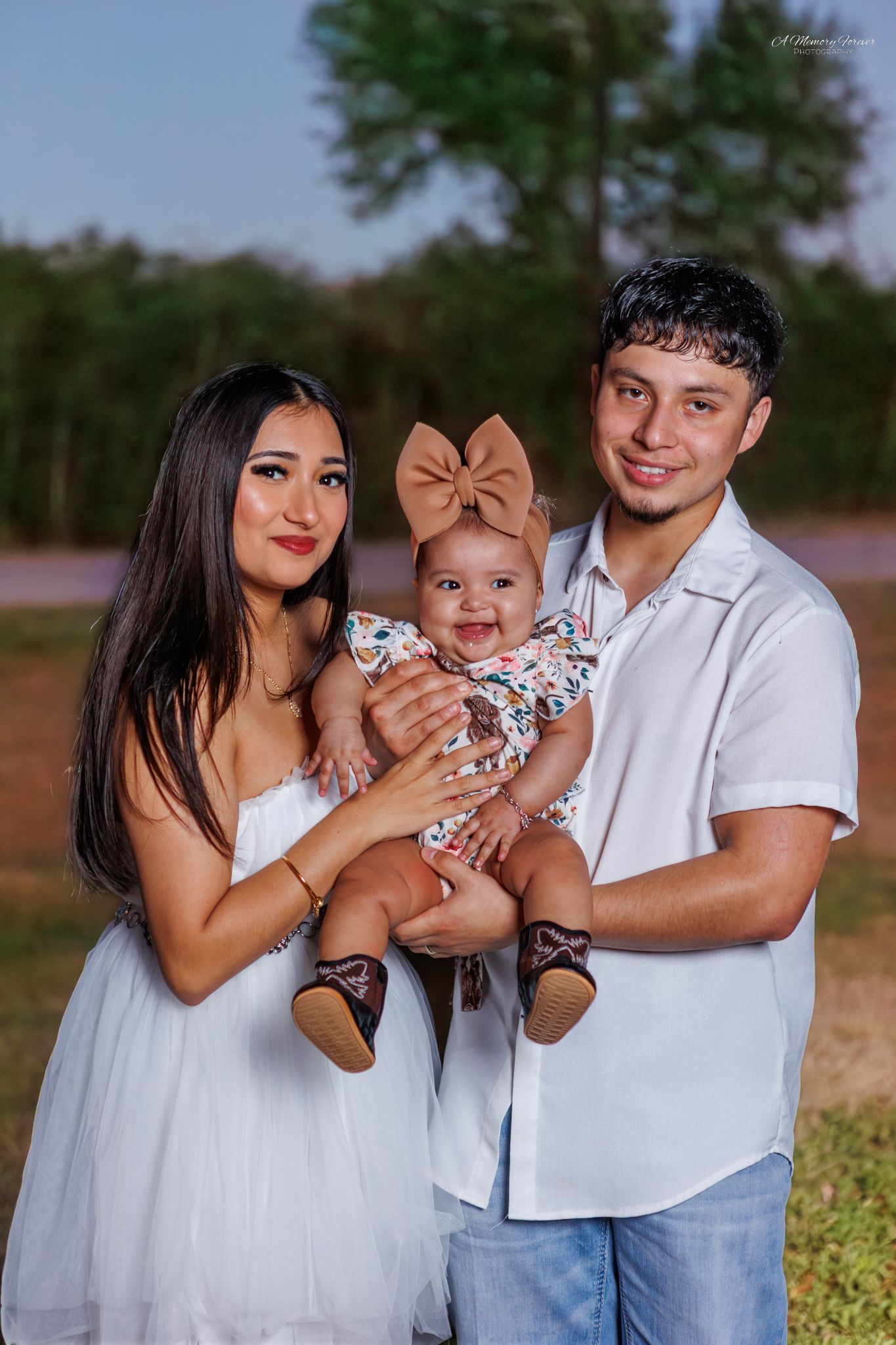 Young Hispanic family poses outdoors, parents in white holding smiling baby girl with large bow headband.