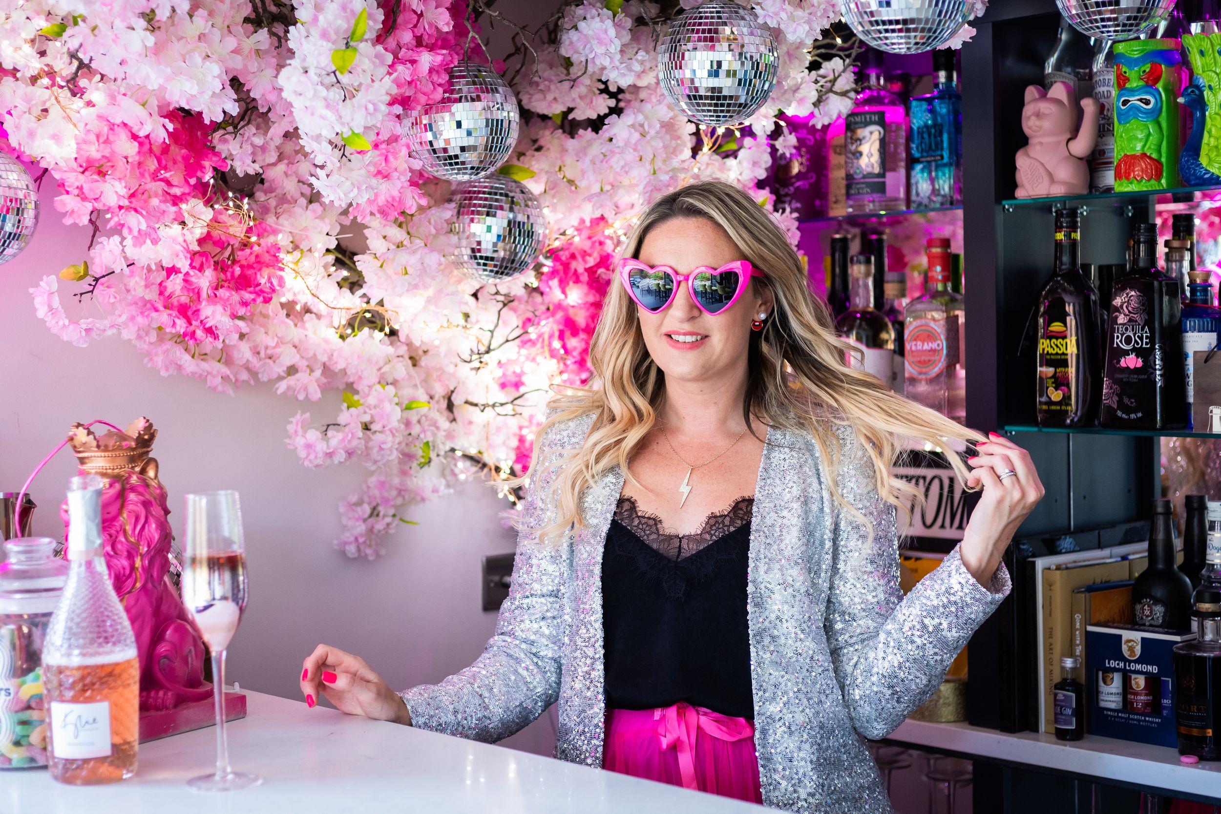 Woman in sunglasses and blazer poses at a pink floral bar with champagne and disco balls.