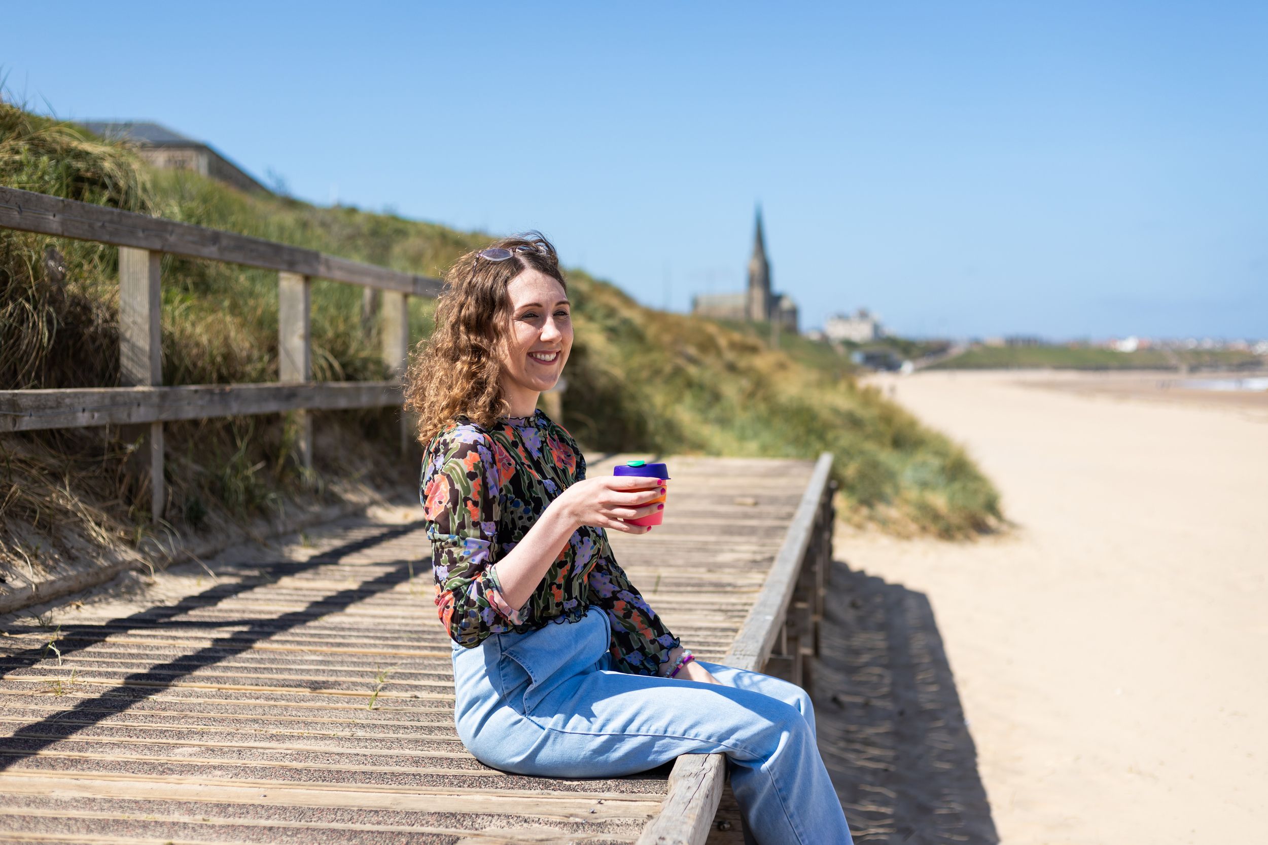 Person in denim sitting on wooden boardwalk at beach with rolling hills and blue sky in background.
