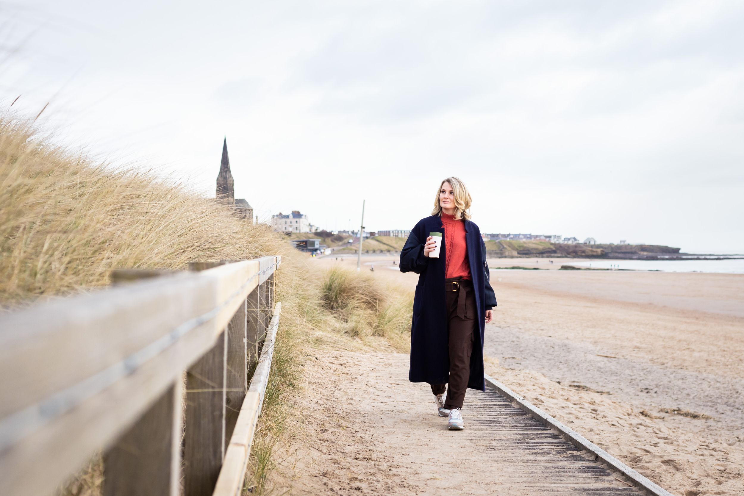 Woman in navy coat walks on beach boardwalk near coastal town with church spire on overcast day.