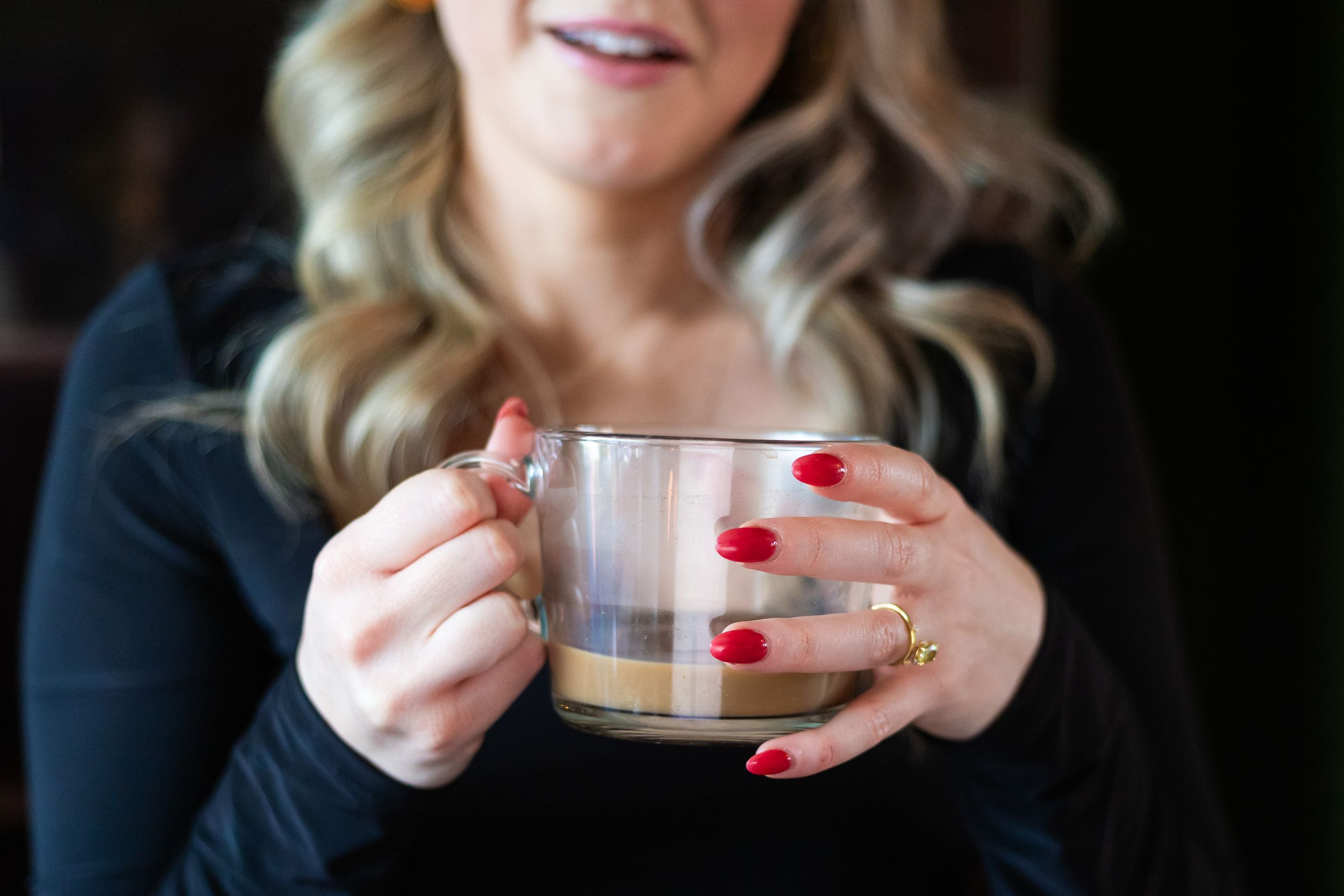 Woman with red nails and blonde hair holding a clear glass coffee cup in a cozy cafe setting.