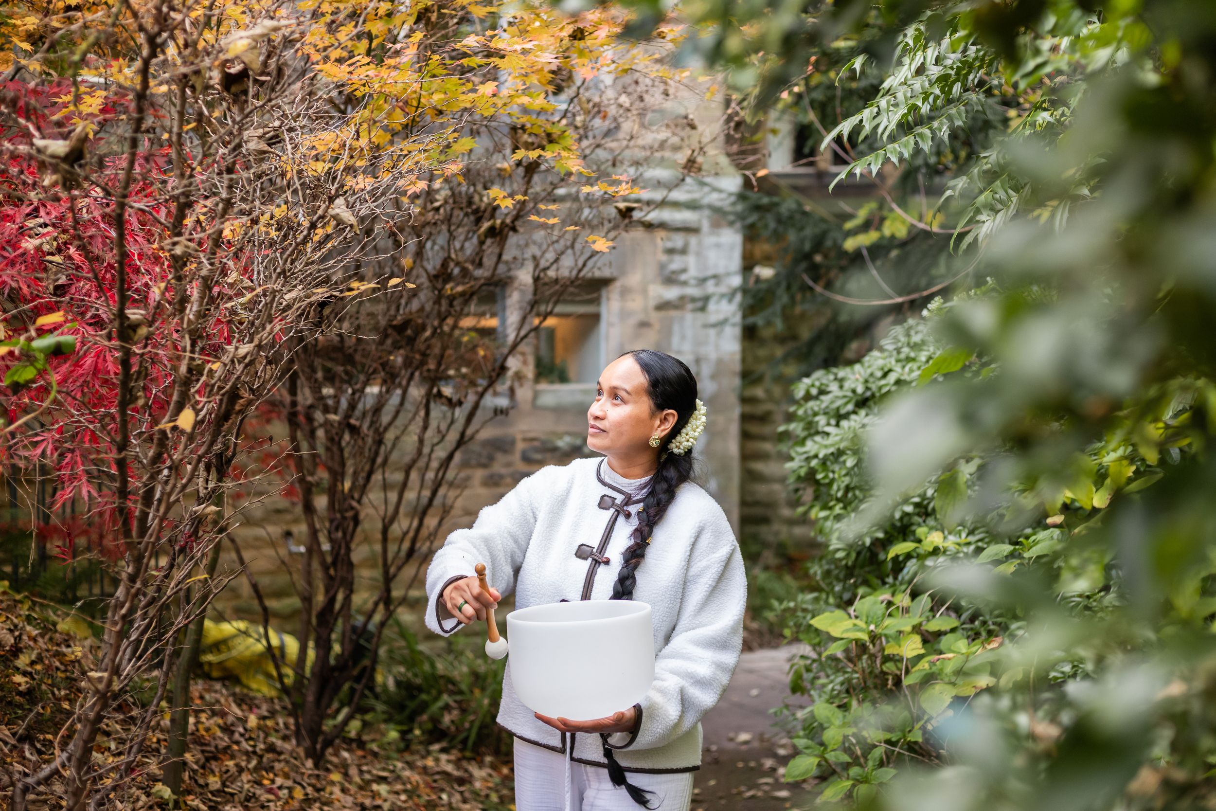 A figure in a white hoodie stands in a garden with autumn foliage and stone walls in the background.