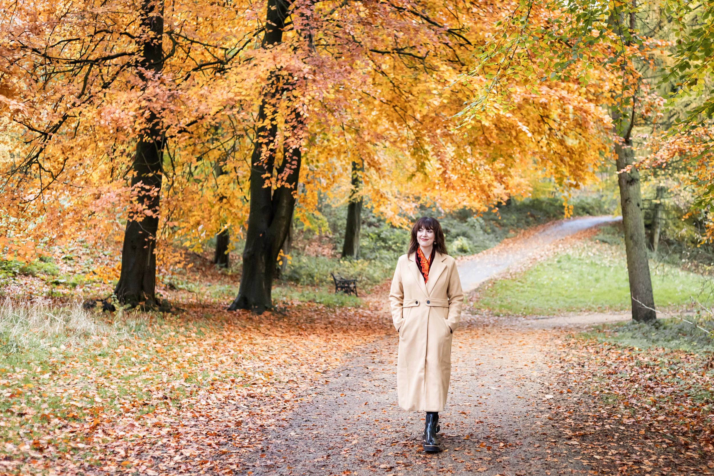 Woman in beige coat walking on a leaf-covered path surrounded by vibrant autumn trees in a park.