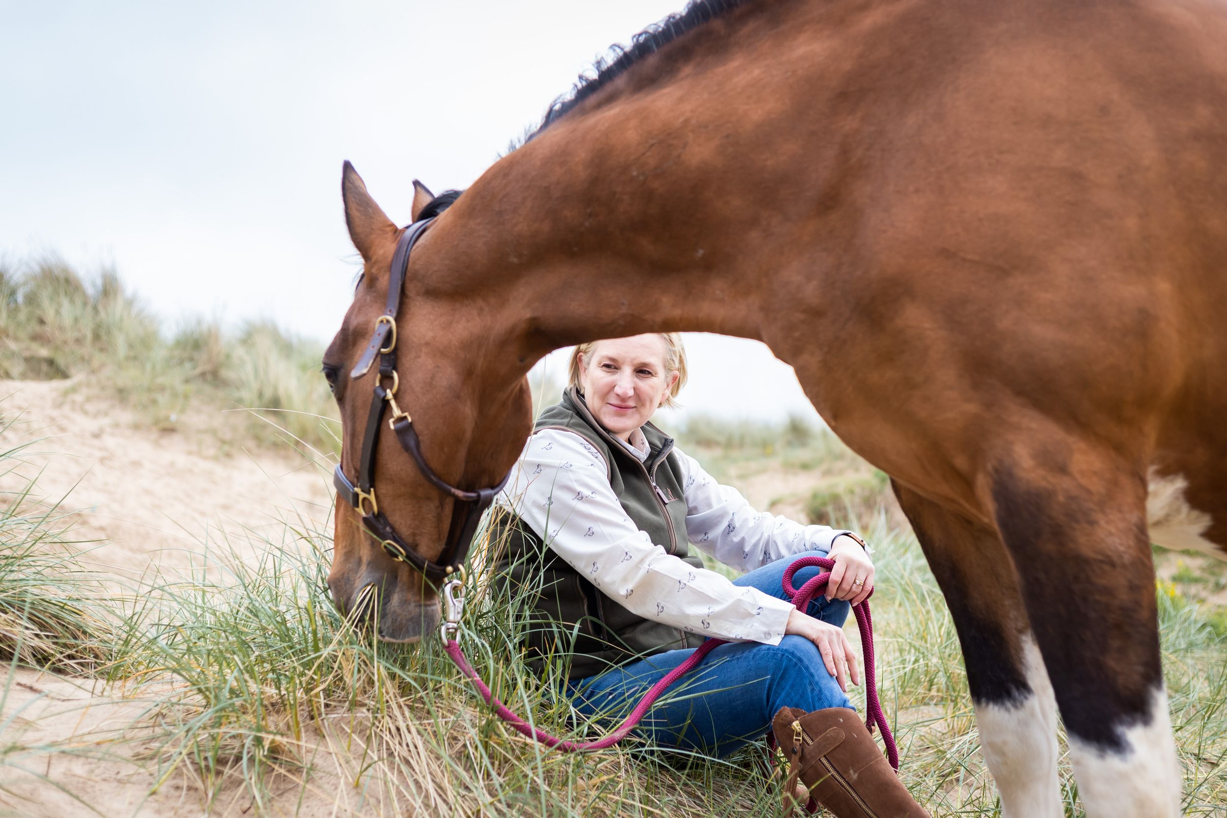 Woman sitting on sandy dunes smiling beside a brown horse grazing on grass near the beach.