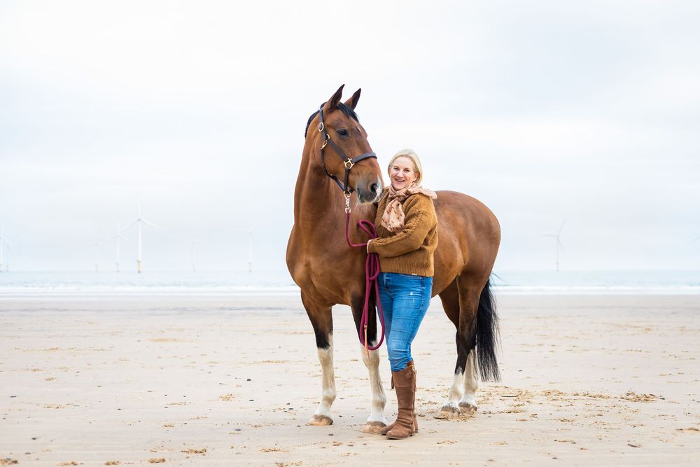 A beautiful brown horse walks along a sandy beach with someone in blue clothing on a cloudy day.