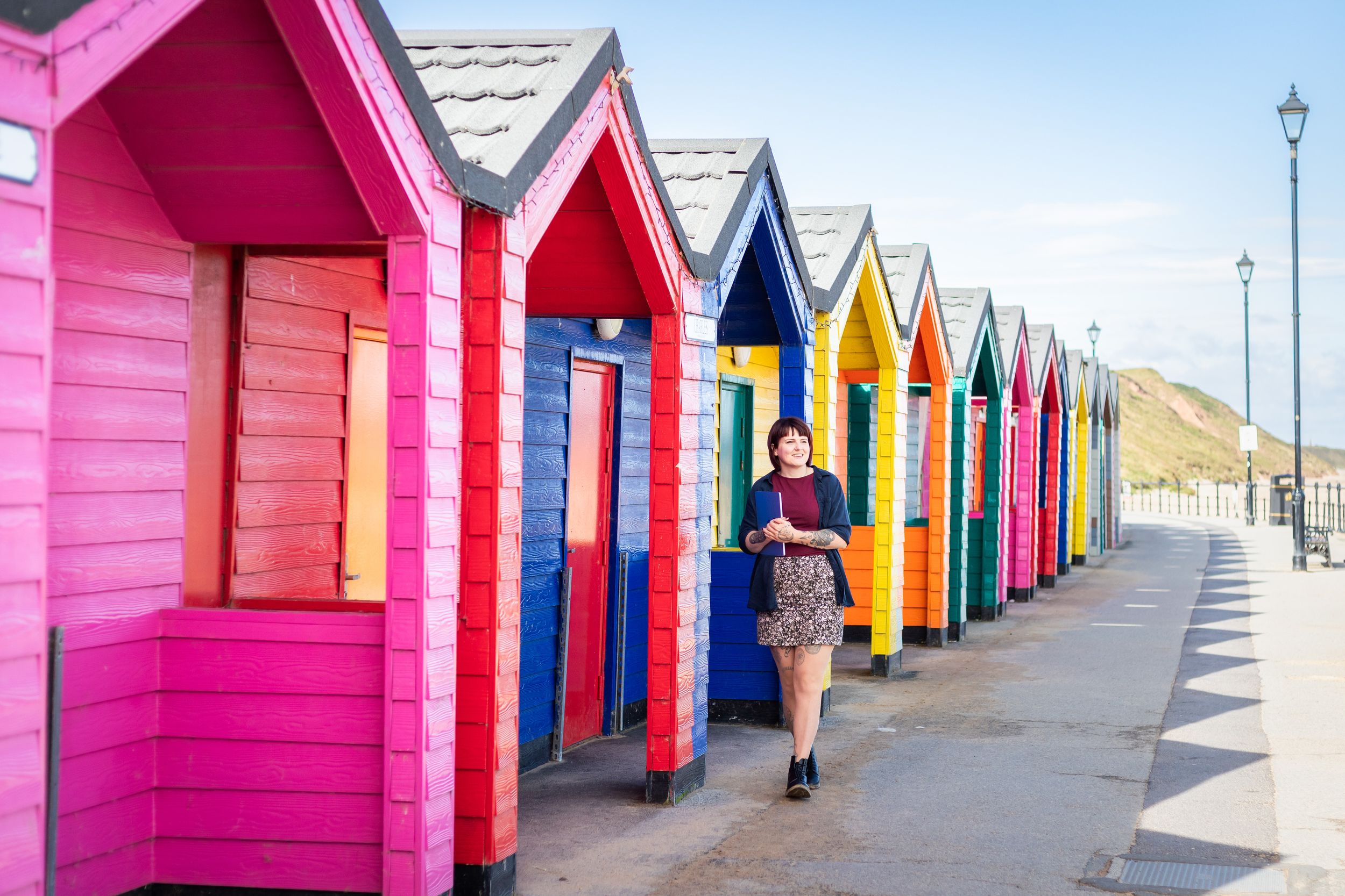 Woman strolling past vibrant multicolored beach huts in a row on a sunny seaside promenade.