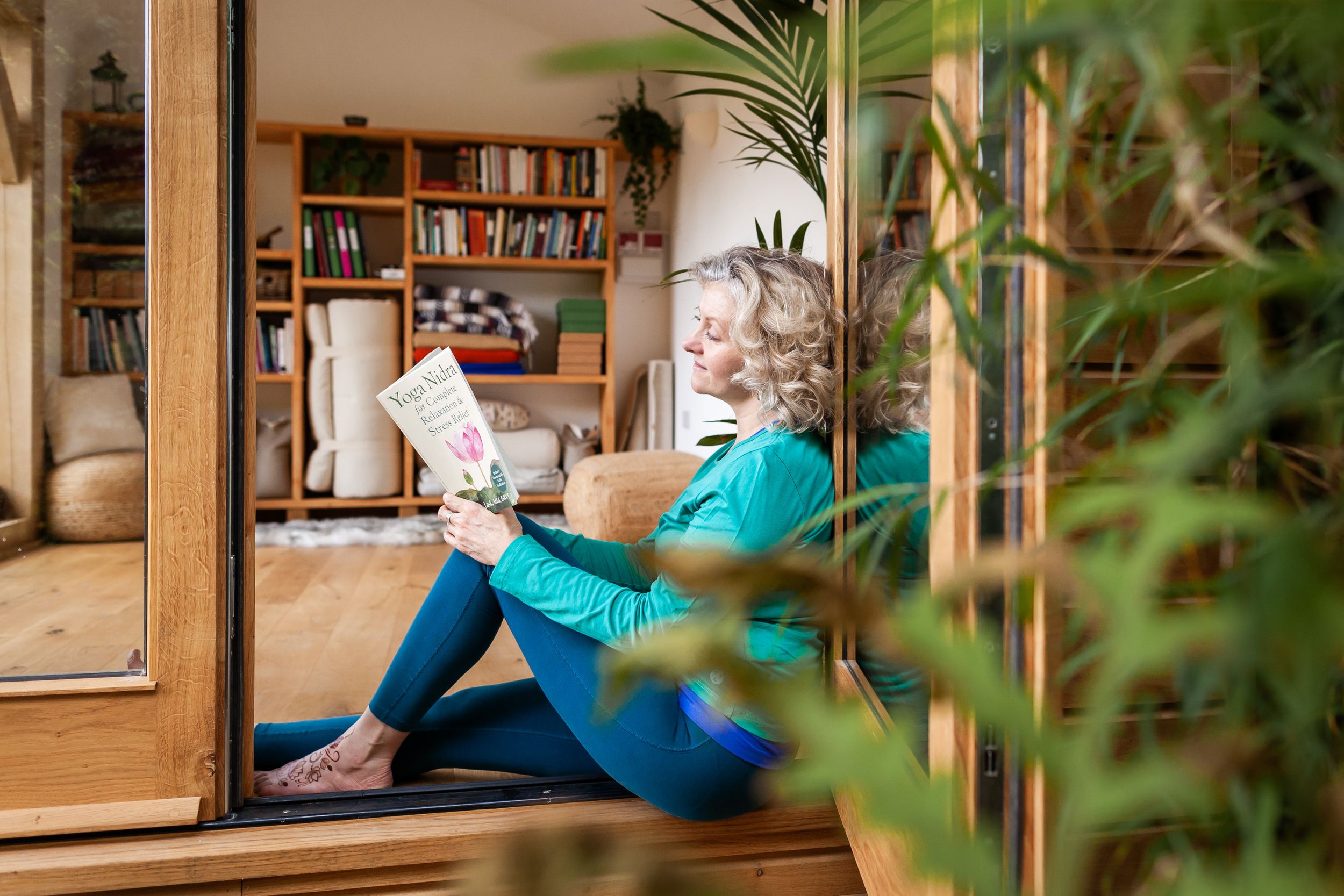 Middle-aged woman relaxing indoors, reading a magazine while seated by wooden doors surrounded by lush green plants.