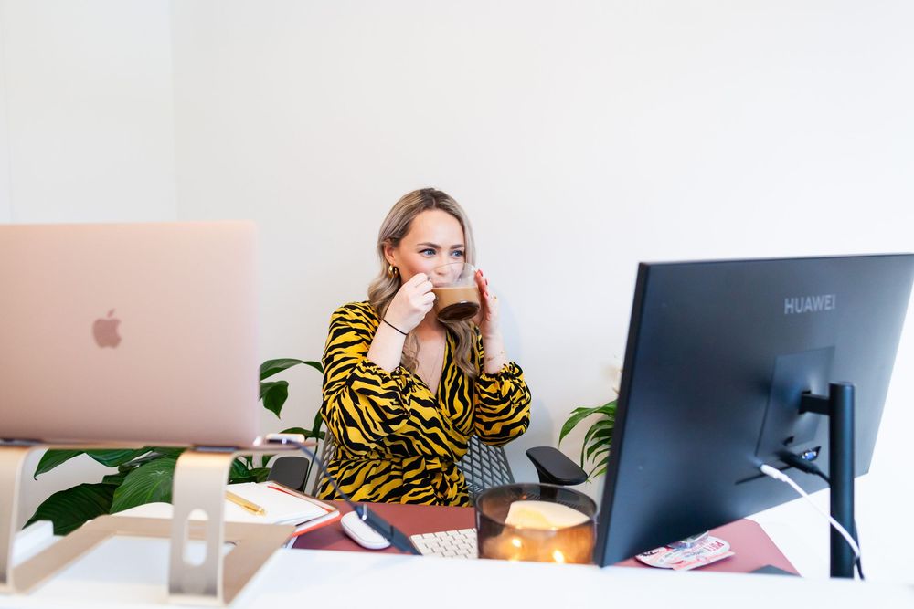 Remote worker in yellow and black patterned sweater sits at desk with computer monitor and houseplant in background.