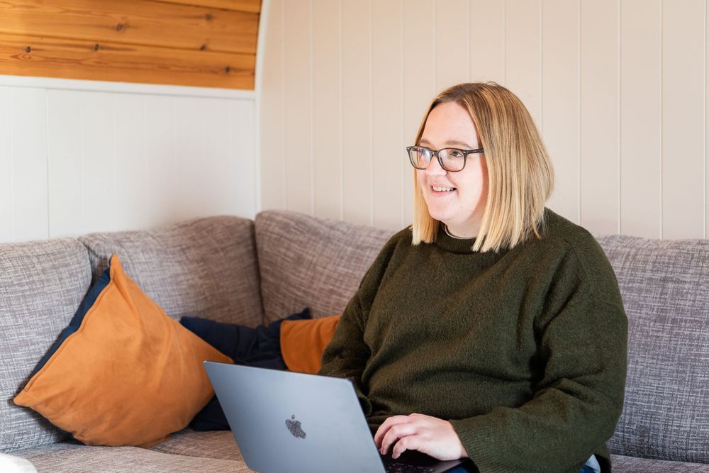 Someone in a dark green sweater works on a MacBook laptop while sitting on a gray couch with orange throw pillows.
