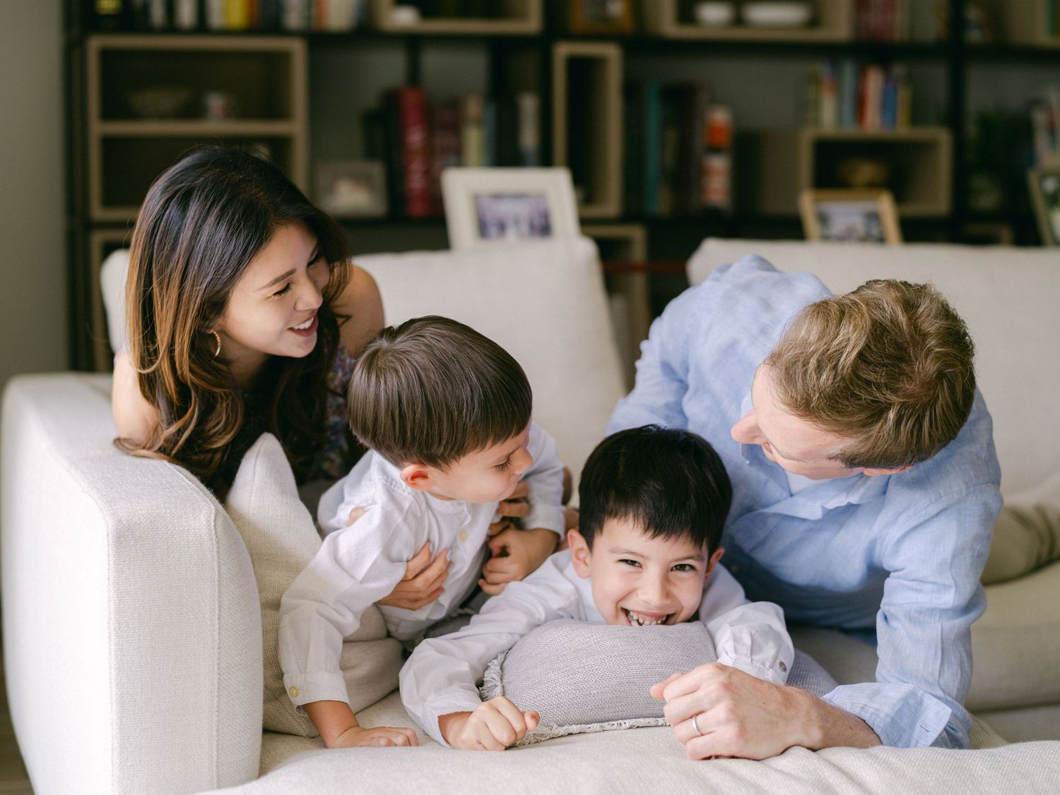 Family of four laughing and playing together on a white sofa in a cozy living room with bookshelves.