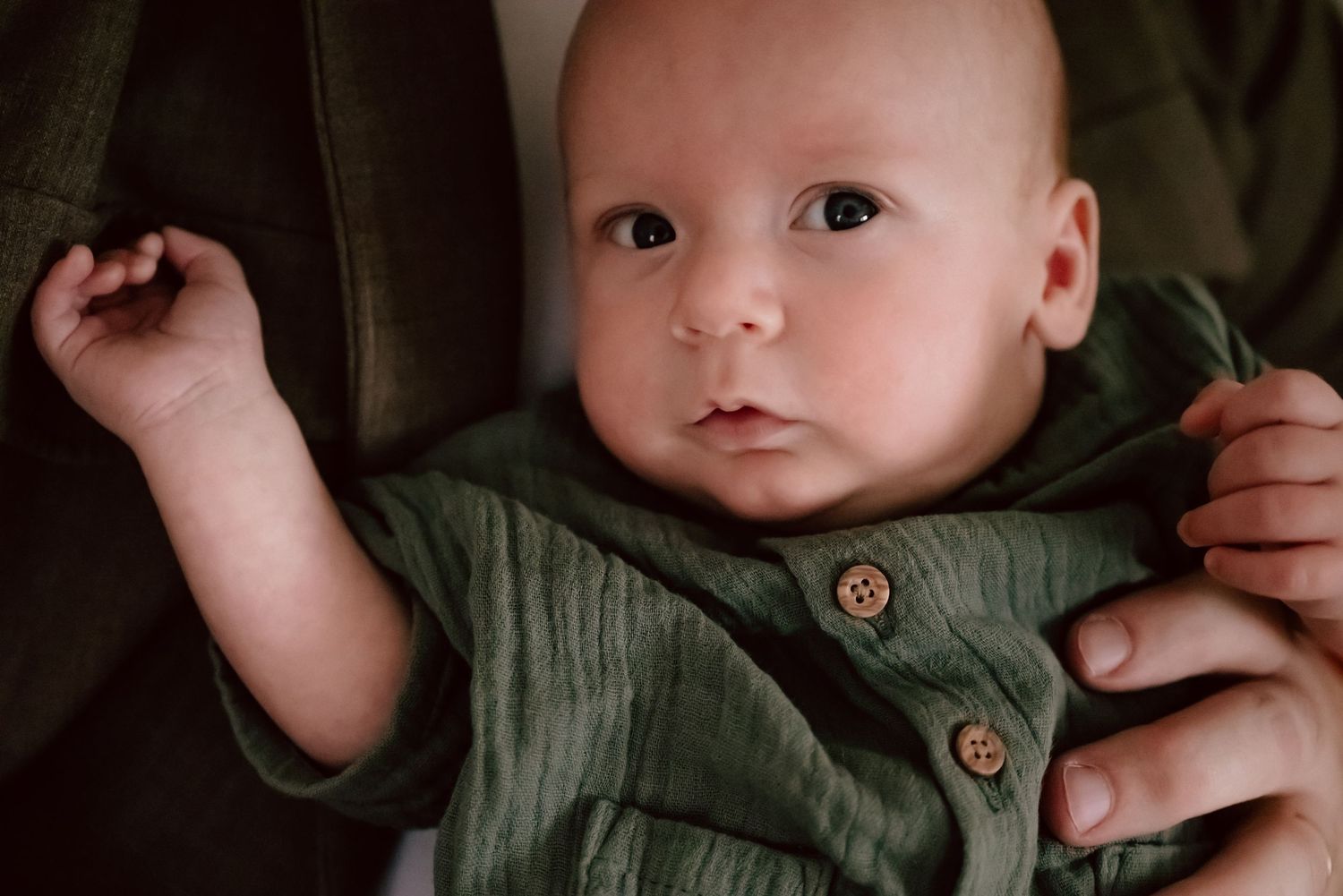 Baby wearing an olive green button-up outfit lying on a dark background with a soft, gentle expression.