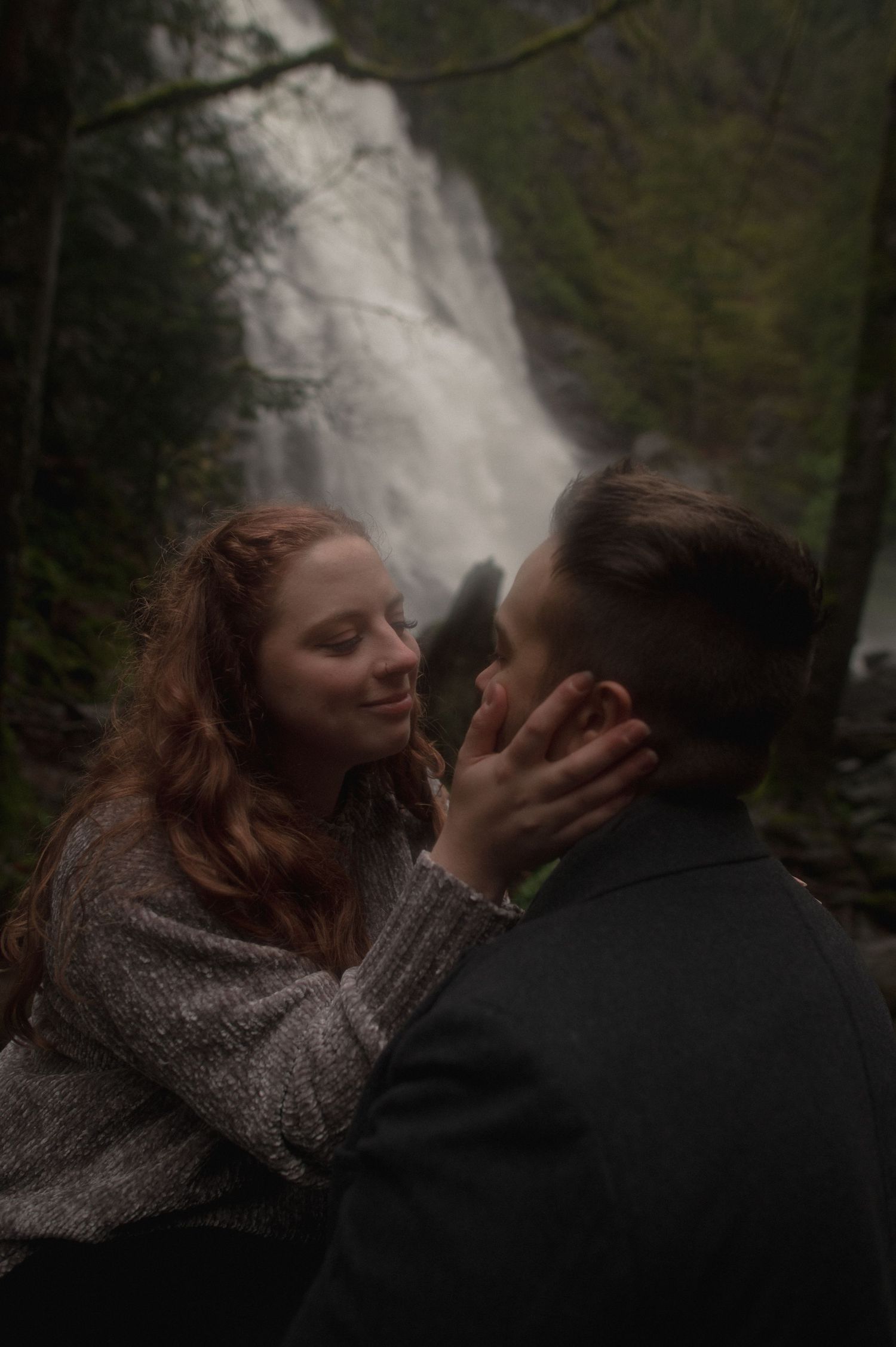 A romantic moment captured in front of a majestic waterfall in a lush forest setting.