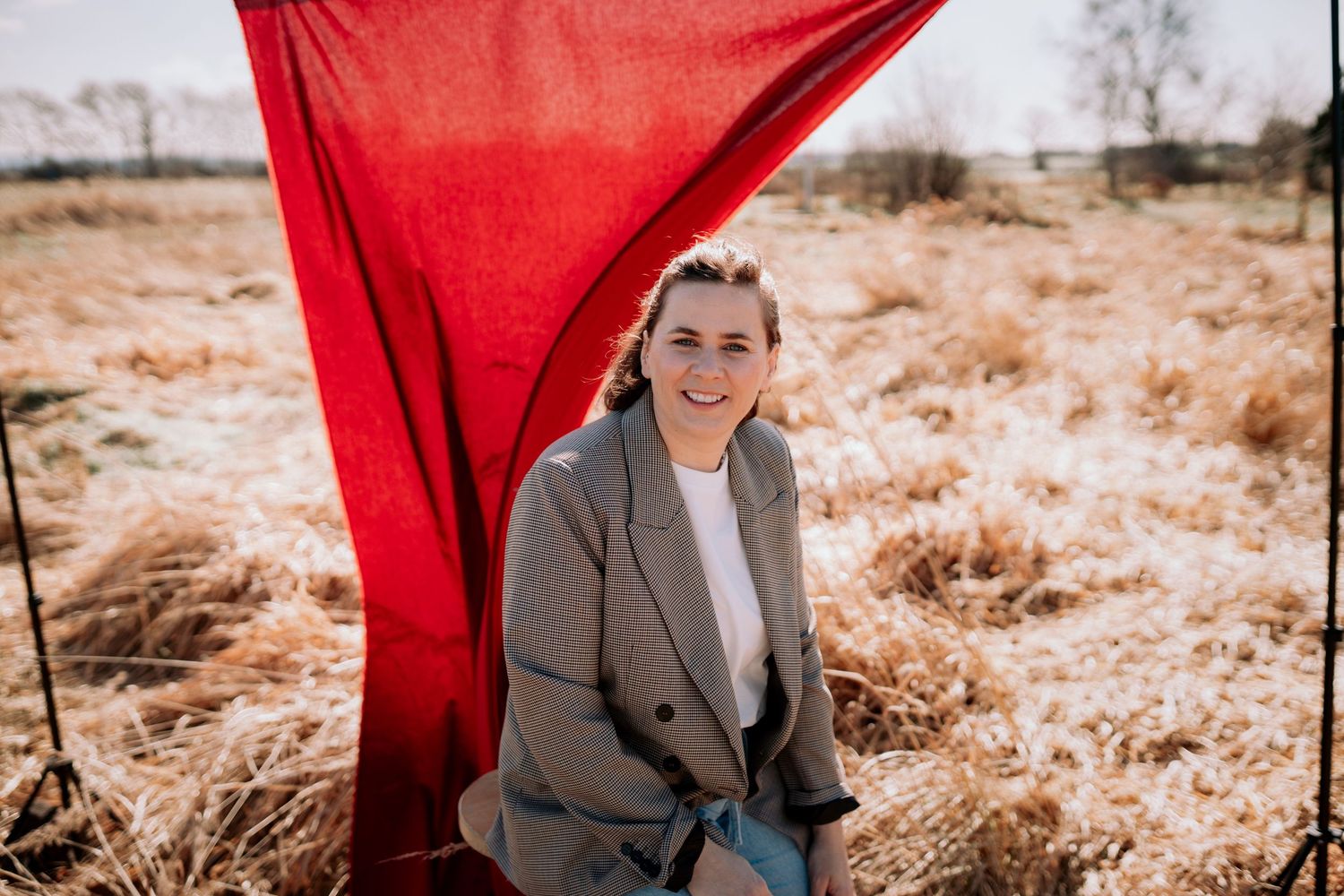 Hey Elli Fotografie sitzt auf einem Stuhl in einem Grasfeld mit einem roten hintergrund und lacht herzlich in die Kamera. Die Sonne scheint.