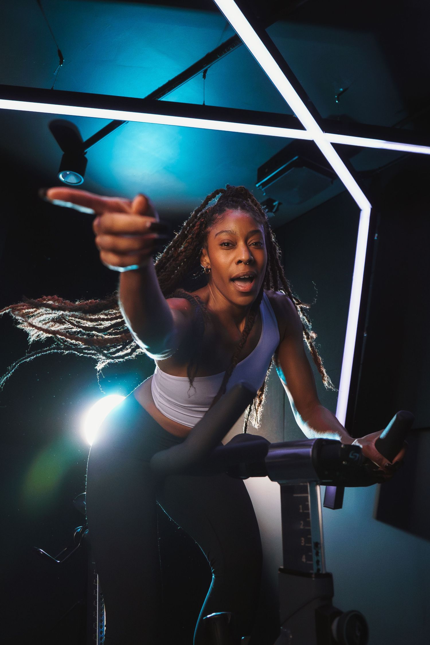Enthusiastic cyclist with braids leading an energetic spin class under blue lighting.
