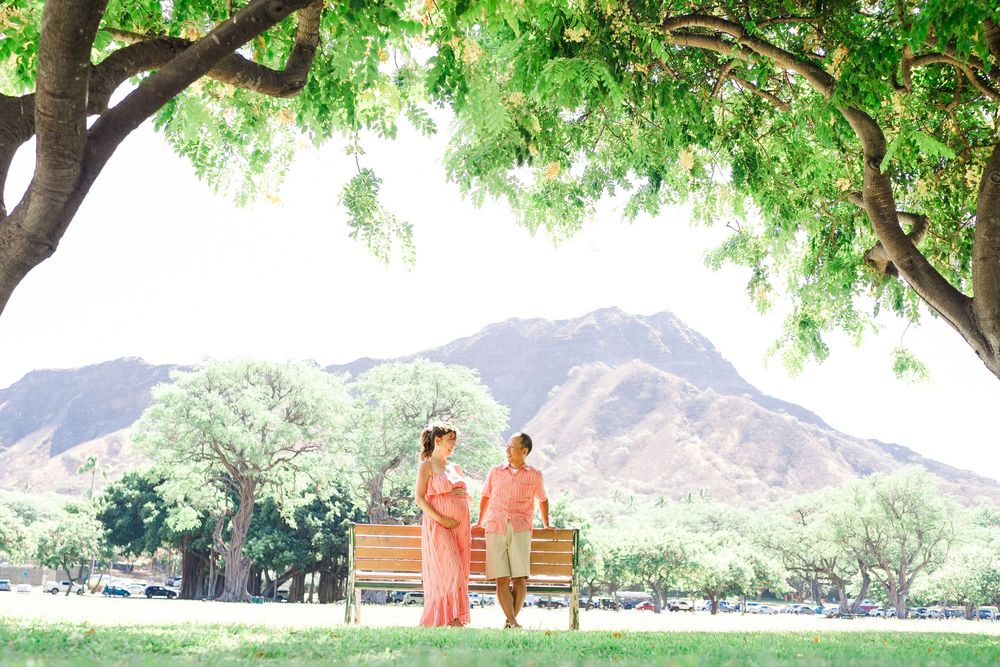 A person in orange robes sits under a large green tree with mountains in the background on a sunny day.
