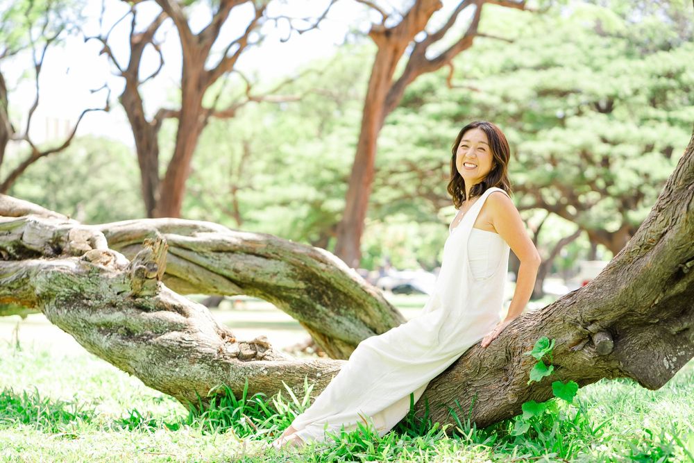Someone in a flowing white dress relaxes against a large tree trunk in a peaceful garden setting.