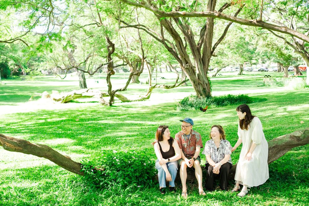 A group relaxes together on a grassy hill under the shade of large trees in a peaceful park setting.