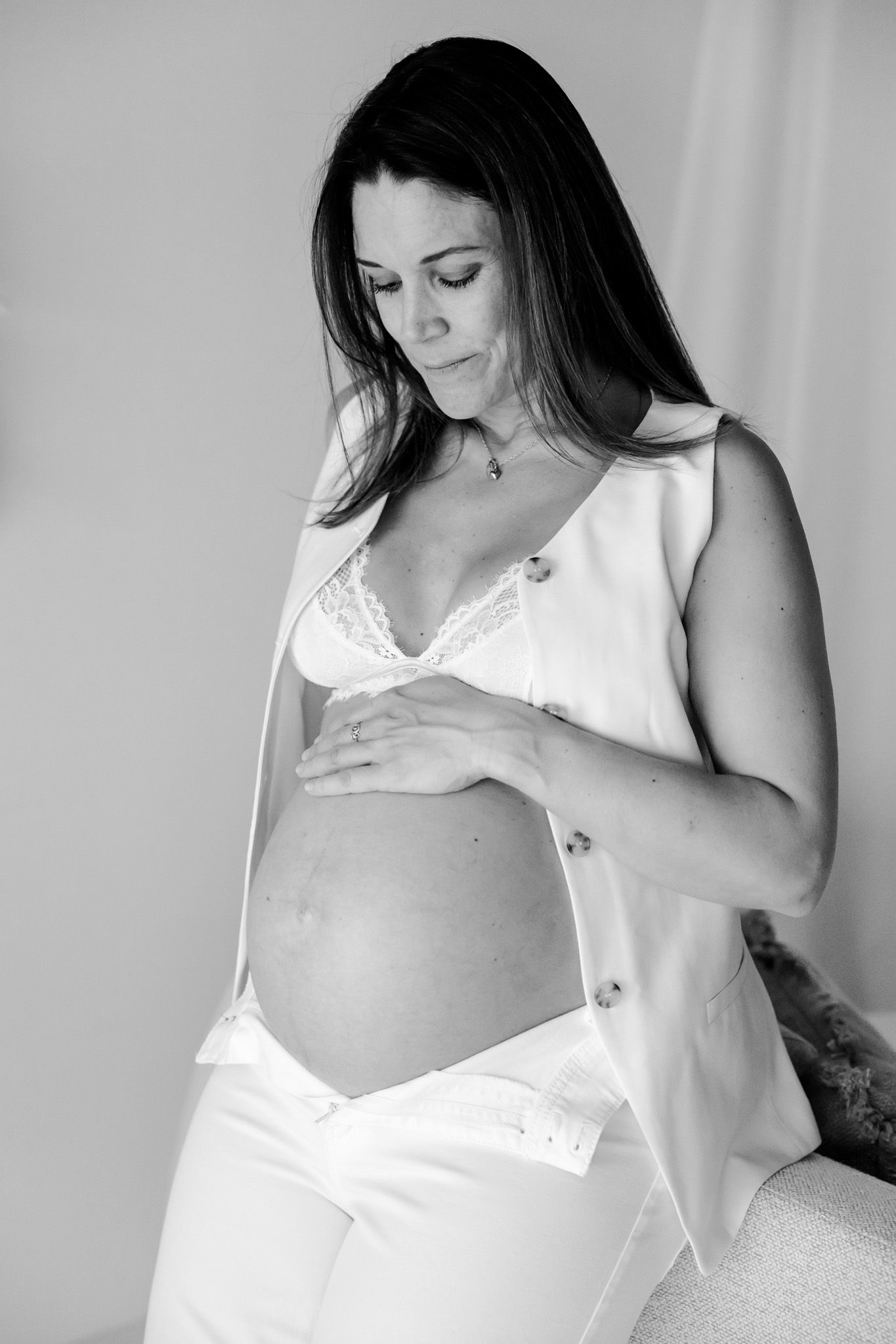 Gravid kvinde i hvid kjole holder sin bare mave med begge hænder mod en lys baggrund. Sorthvid billede. Fotograf Middelfart