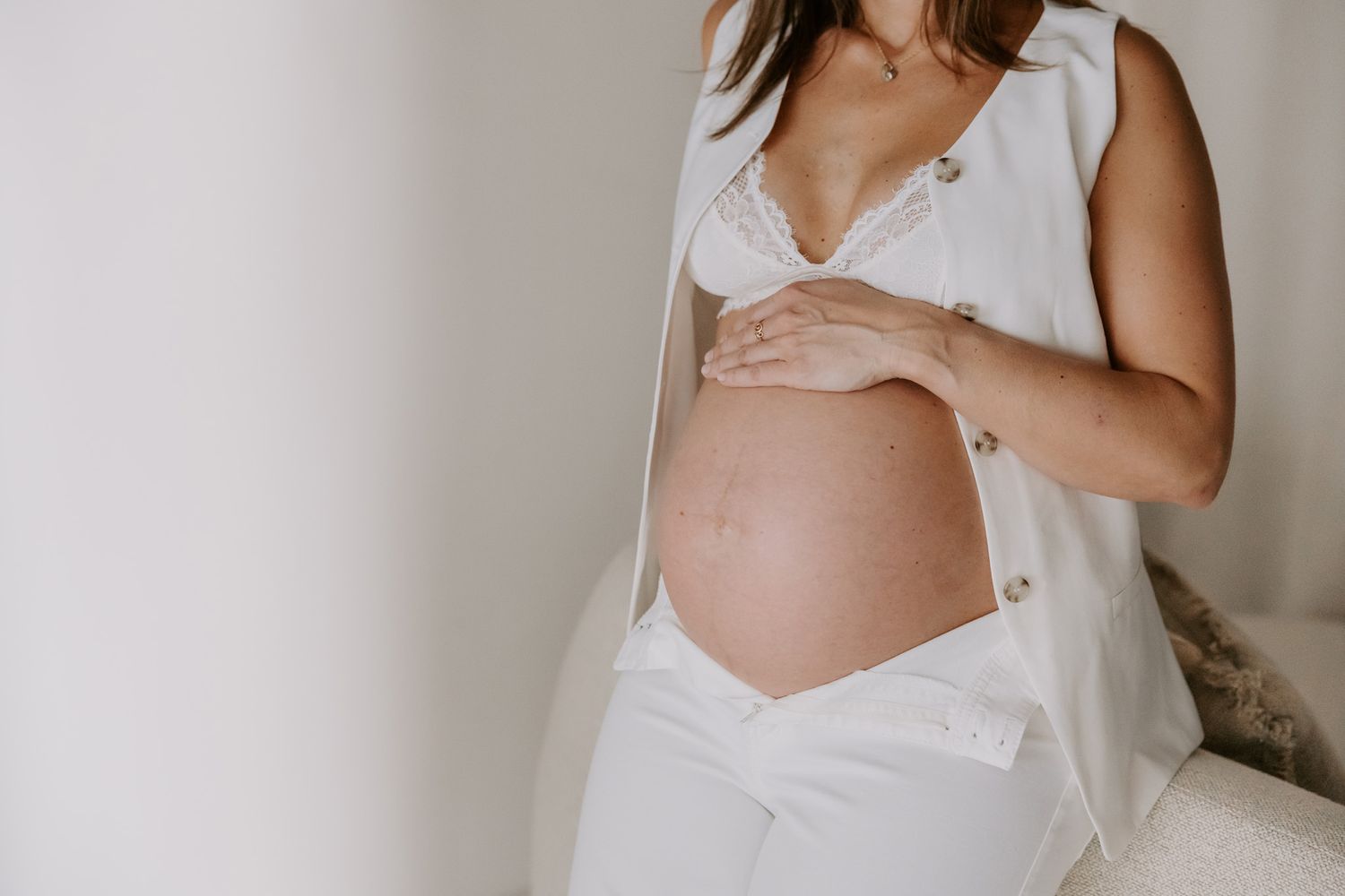 Gravid kvinde i hvid kjole holder sin bare mave med begge hænder mod en lys baggrund. Fotograf Middelfart