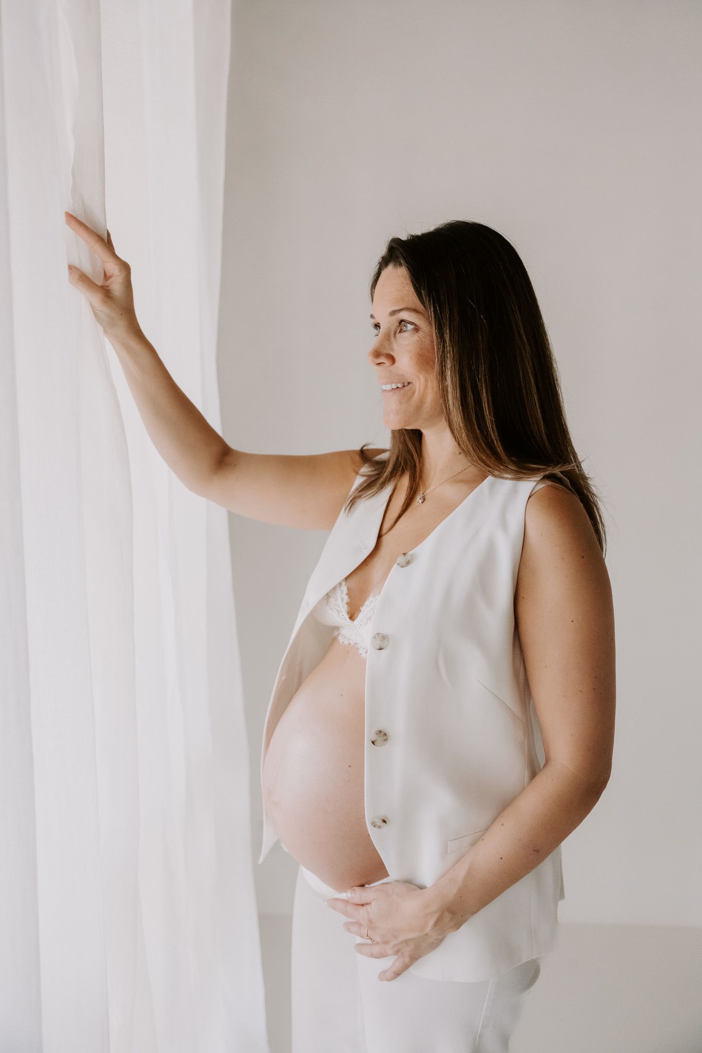 Gravid kvinde i hvid kjole holder sin bare mave med begge hænder mod en lys baggrund. Fotograf Middelfart