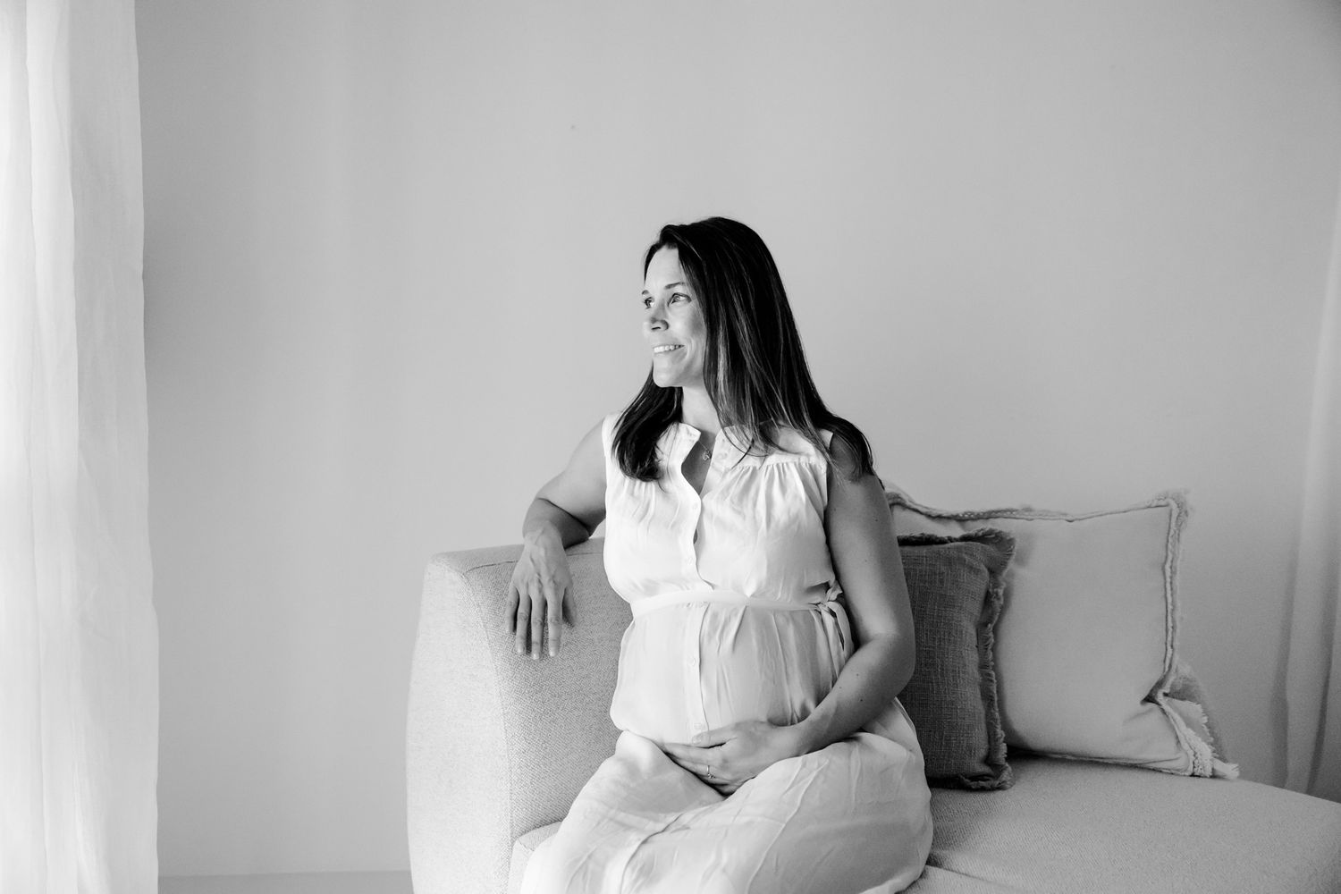 Sort-hvidt billede af en smilende gravid kvinde, siddende på en sofa og holder om maven i et lyst rum. Fotograf Middelfart