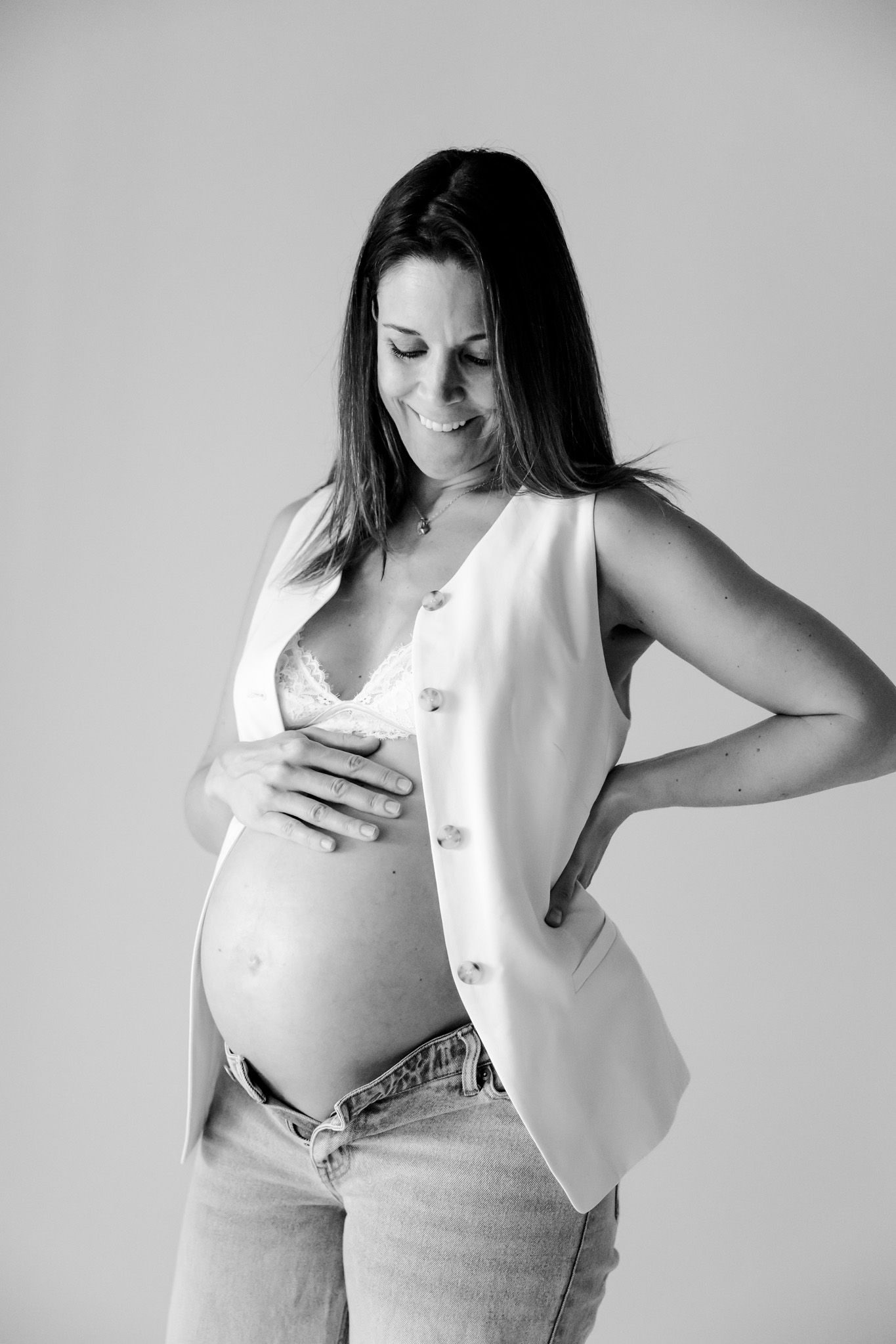 Sort-hvidt billede af en smilende gravid kvinde i jeans og vest, der holder om sin bare mave. Fotograf Middelfart.