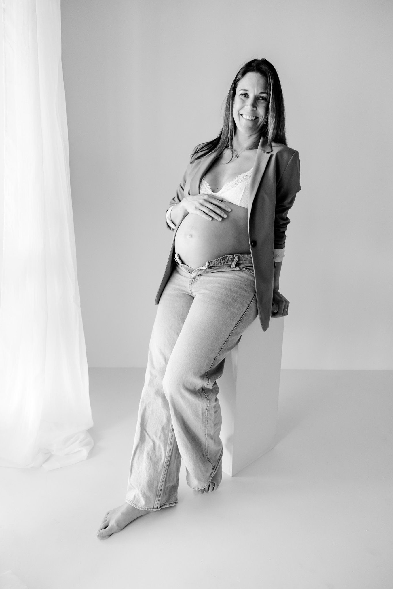 Gravid kvinde i hvid blazer og jeans holder om maven, stående ved vinduet i et lyst, minimalistisk rum. Fotograf i Middelfart.