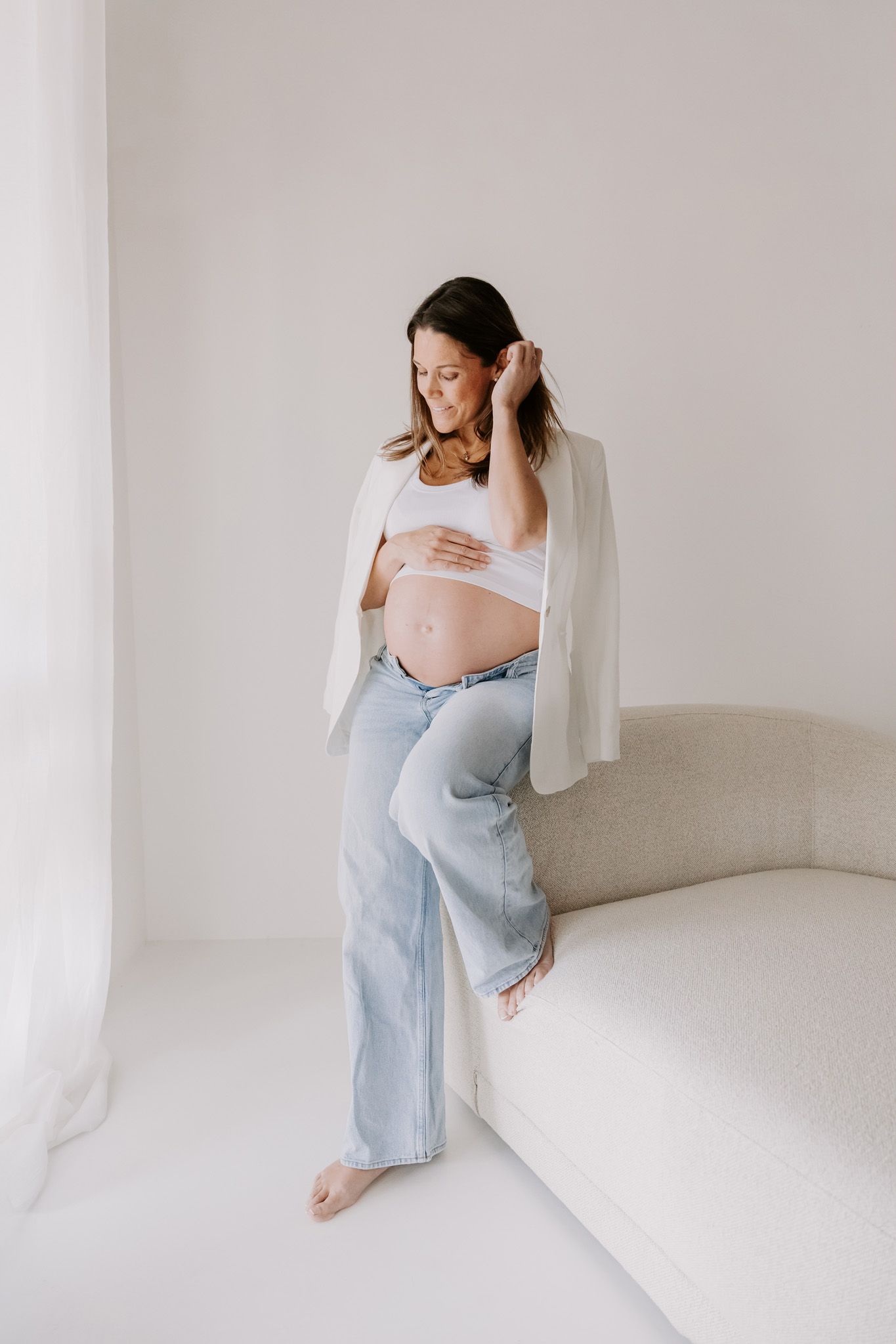 Gravid kvinde i hvid blazer og jeans holder om maven, stående ved vinduet i et lyst, minimalistisk rum. Fotograf i Middelfart.