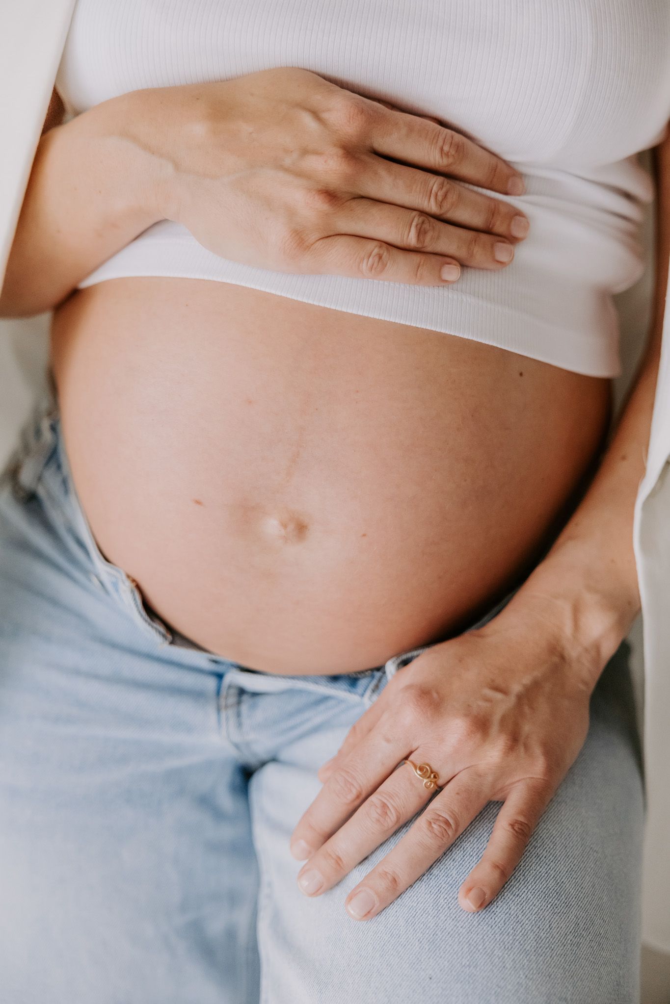 Gravid kvinde i hvid blazer og jeans holder om maven, stående ved vinduet i et lyst, minimalistisk rum. Fotograf i Middelfart.