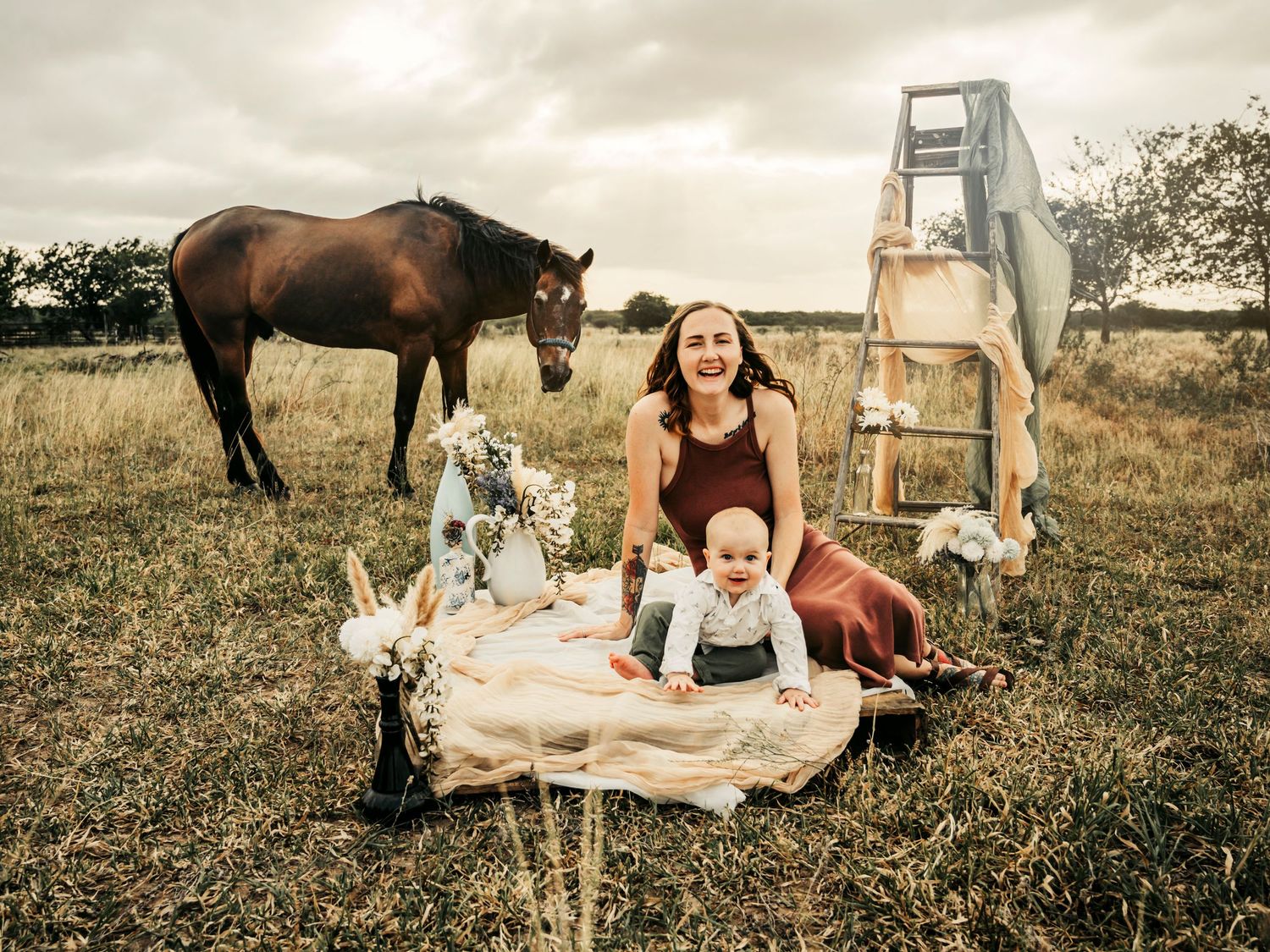 Mother and young son sitting on a blanket in a field, with a horse in the background, enjoying a peaceful moment together.
