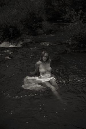A moody black and white photo captures a figure sitting on a rock in flowing water amongst a forested landscape.