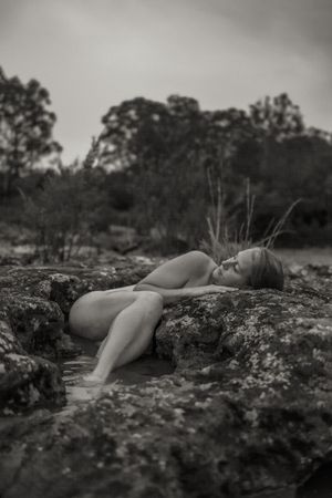 A black and white artistic photograph of a person lying on rugged rocks in nature with trees in the background.