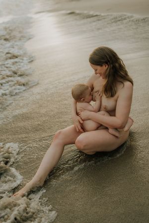 Mother and baby share intimate bonding moment while sitting on sandy beach as waves roll in.