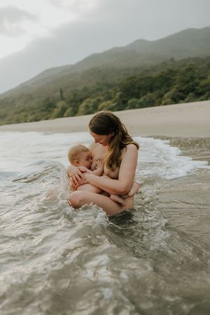 Parent and baby sharing an intimate moment while sitting in shallow ocean waves near a mountainous beach.