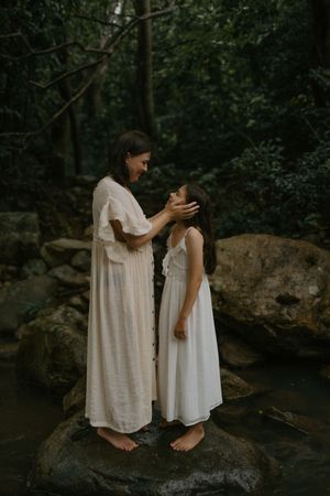 A woman in a white dress tenderly holds a girl's face while standing on rocks in a lush forest setting.