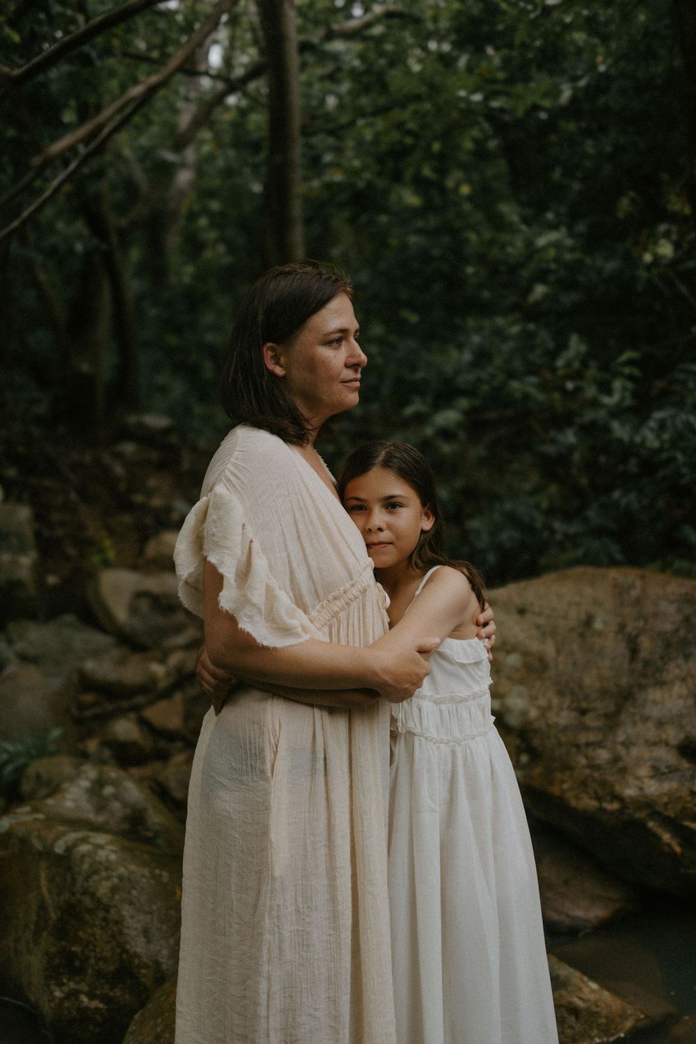 A mother and daughter in white dresses embrace in a lush forest setting, sharing a tender moment.