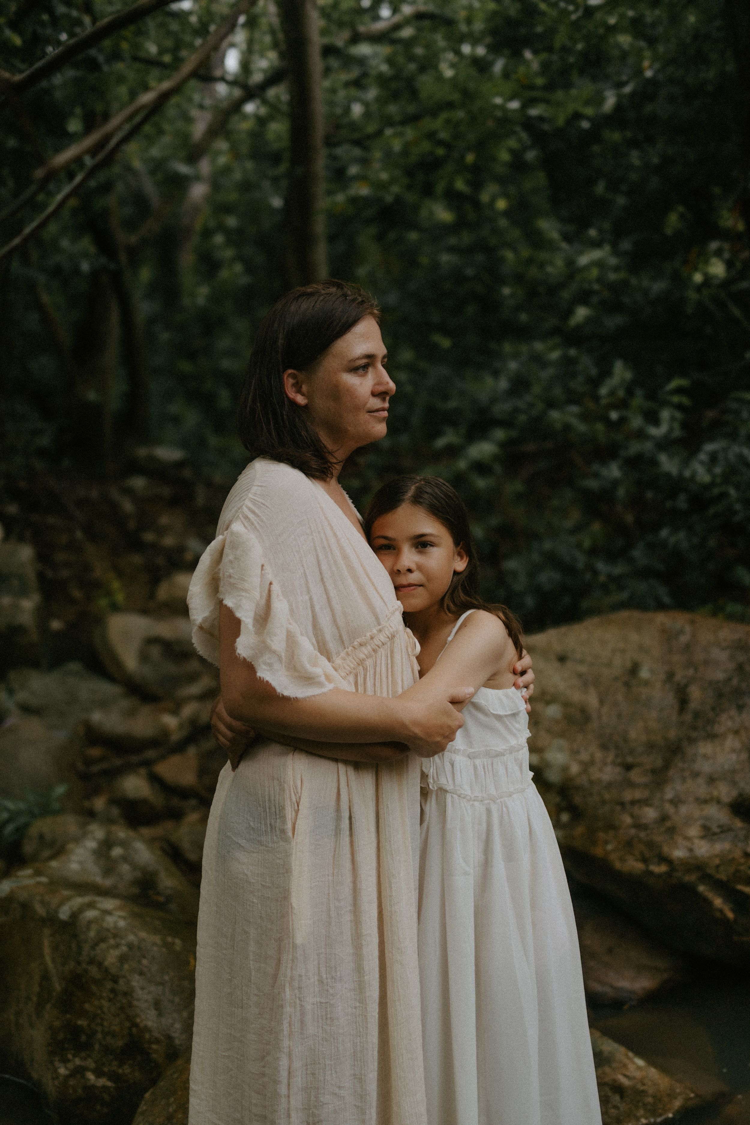 A mother and daughter in white dresses embrace in a lush forest setting, sharing a tender moment.