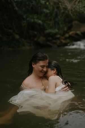 Mother and child share tender embrace while wading in a serene forest stream, wearing white dress.