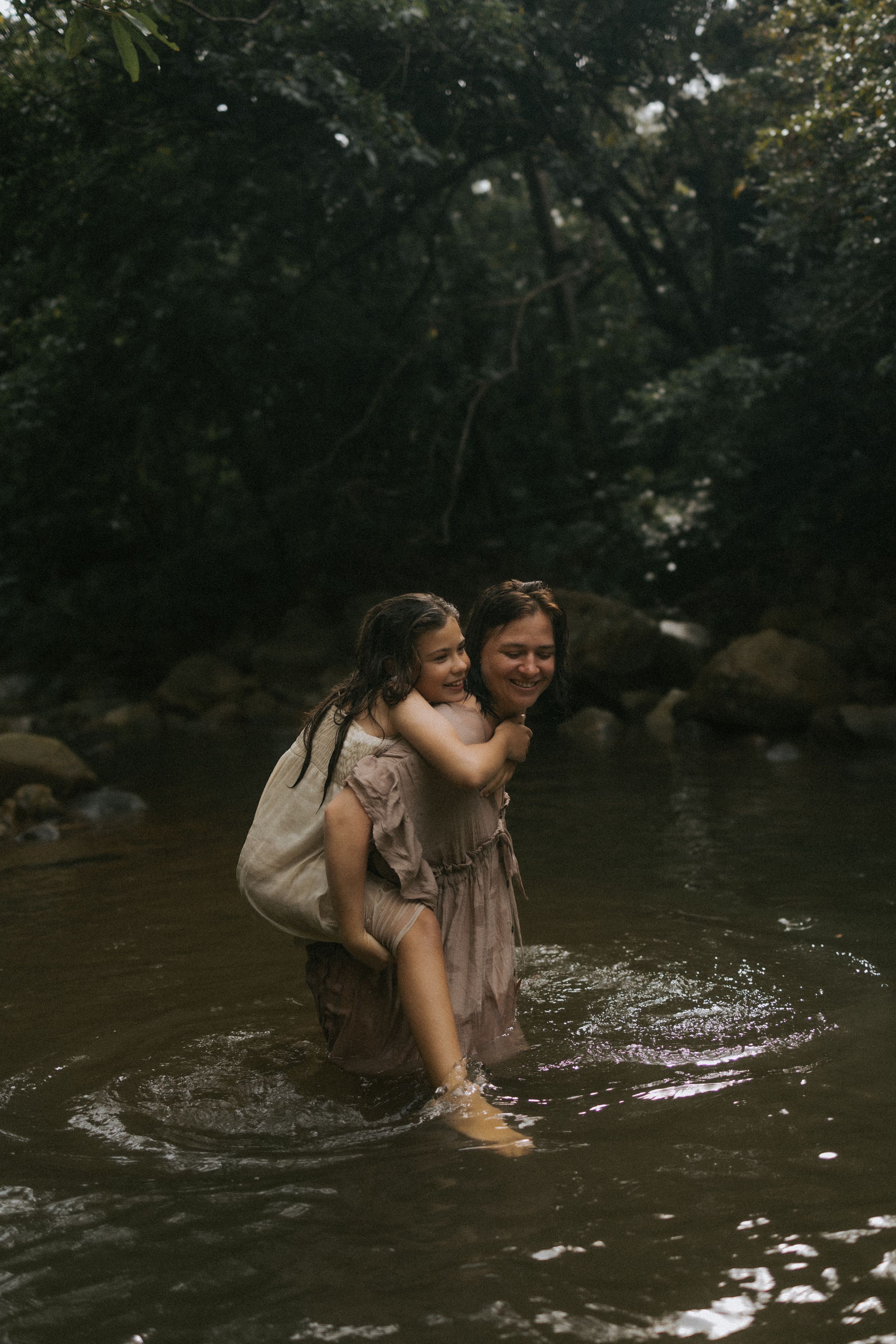 A mother carries her young daughter piggyback while wading through a serene forest river in matching pink dresses.