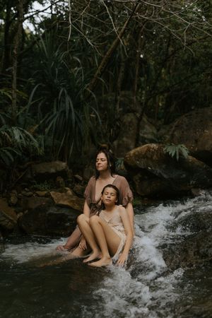 Woman and child sitting peacefully in a flowing forest stream surrounded by lush tropical greenery and rocks.