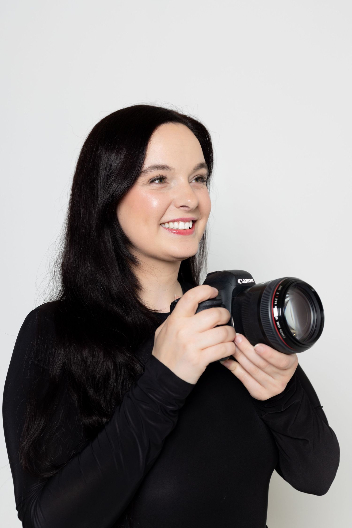 Professional photographer wearing black holding a Canon DSLR camera against white background.