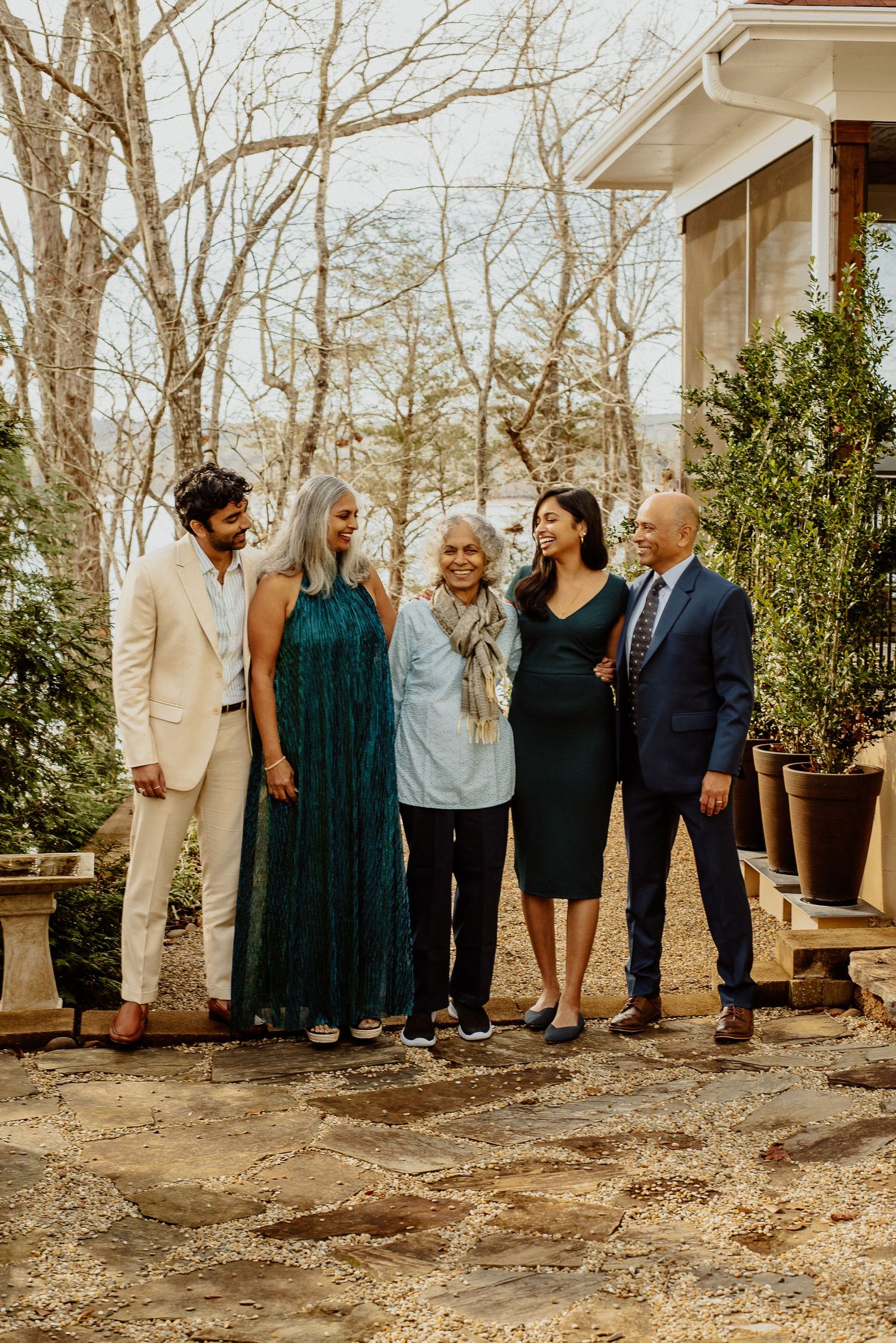 Group in formal attire standing together outside rustic home surrounded by bare trees.
