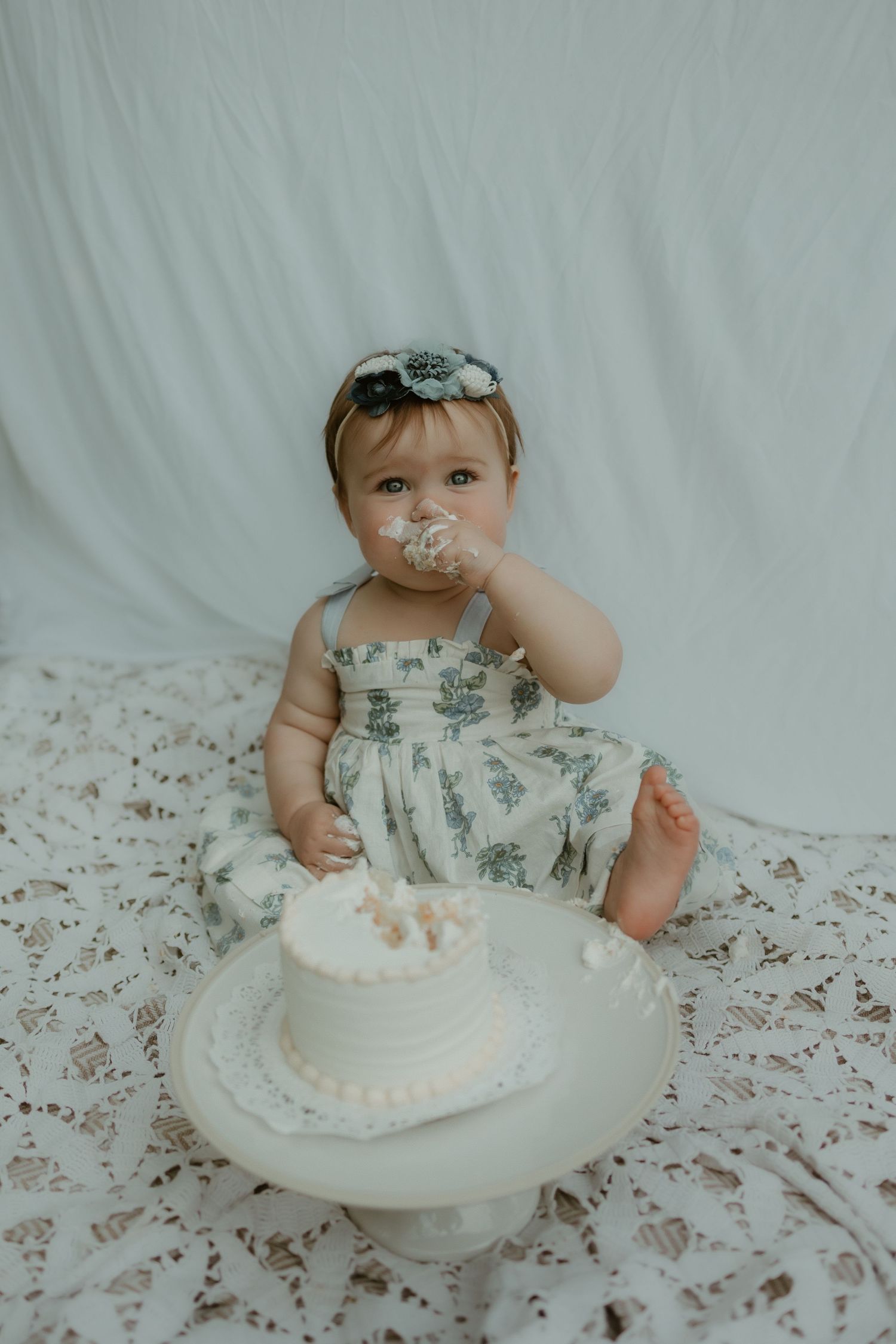 Baby in floral dress sits on white backdrop with cake smash birthday setup and frosting on face during photo session.