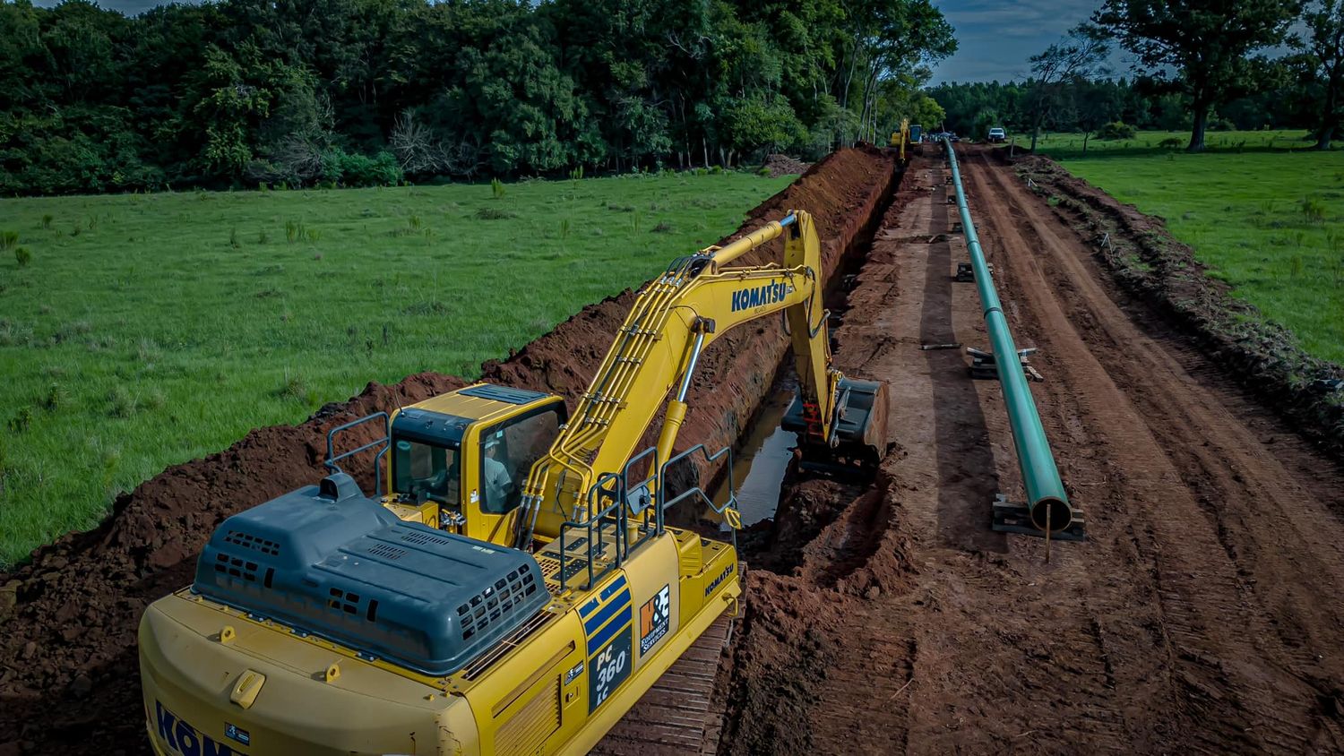 Yellow excavator digging trench for pipeline installation along rural construction site with green fields.