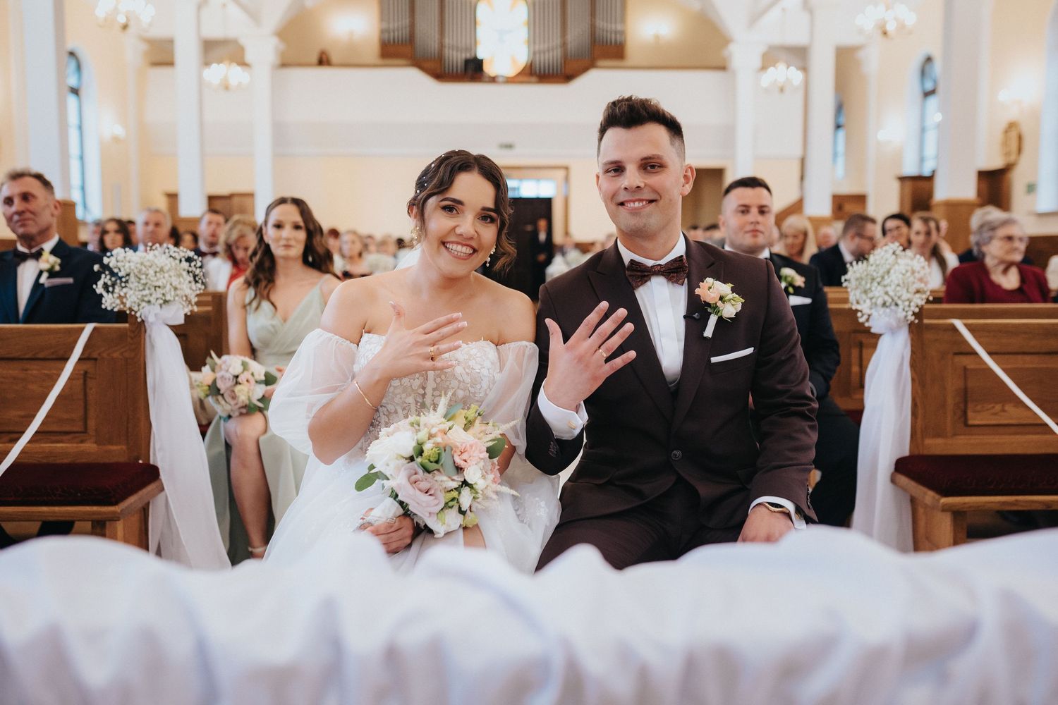 Mlodzi pokazuja obaczki siedzac na krzeslach w kosciele.  Wojciech Grzegorczyk fotograf ślubny