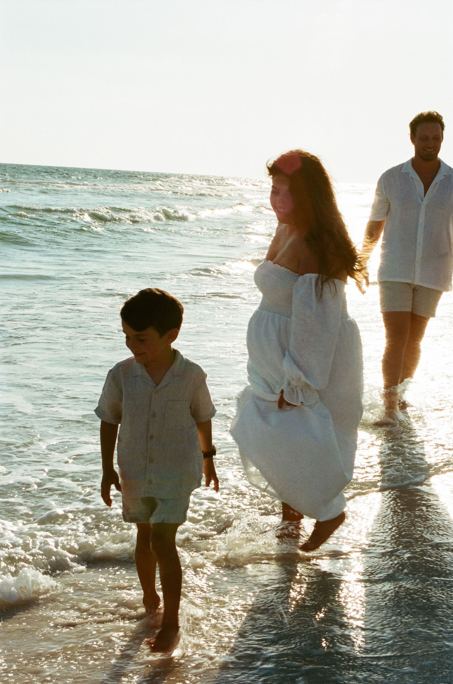 Family wearing white clothing walks together along the beach shoreline at sunset.